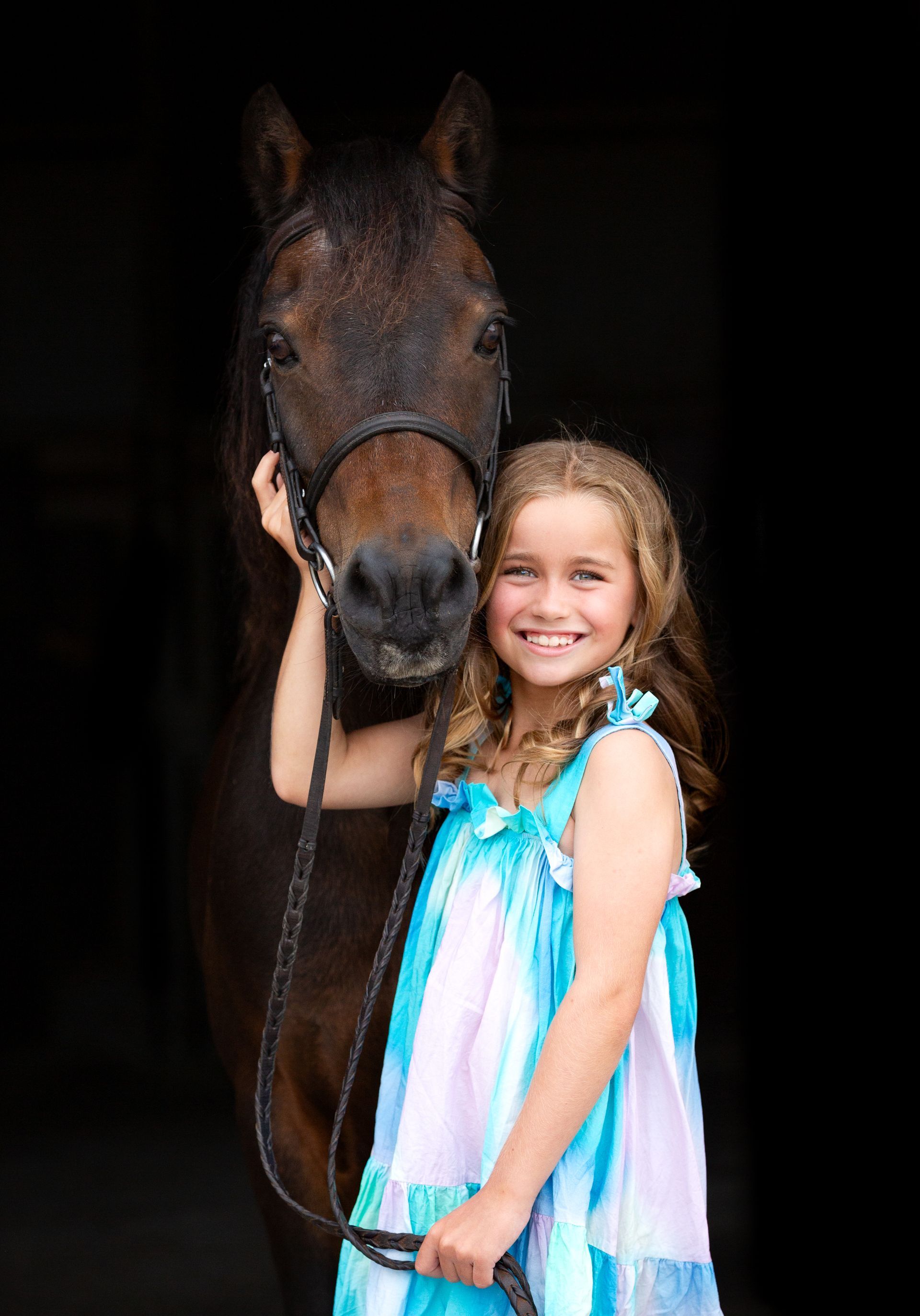 Girl smiles while holding a brown horse, both against a black backdrop.