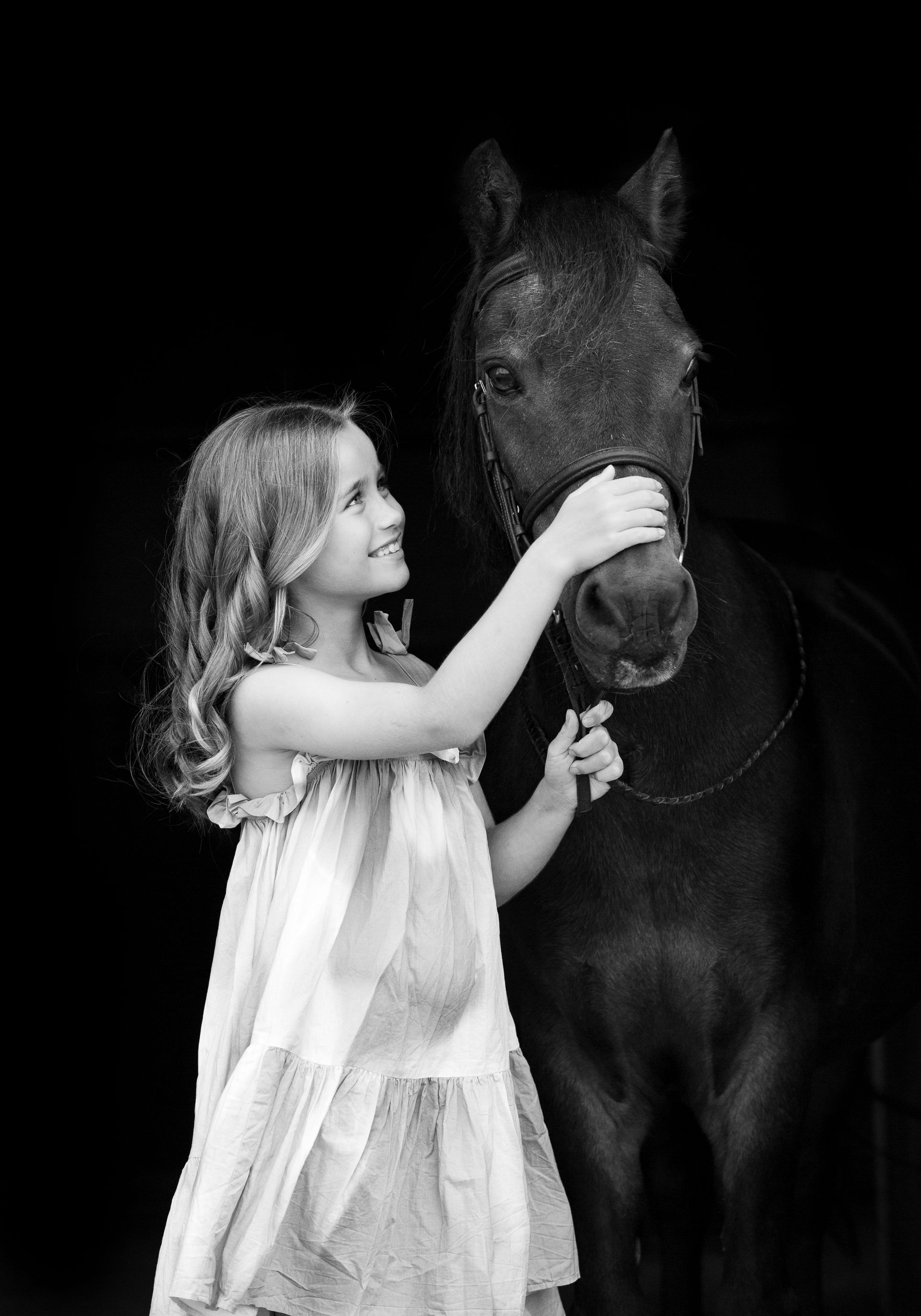 Girl in a dress petting a dark horse, both smiling, against a black background.