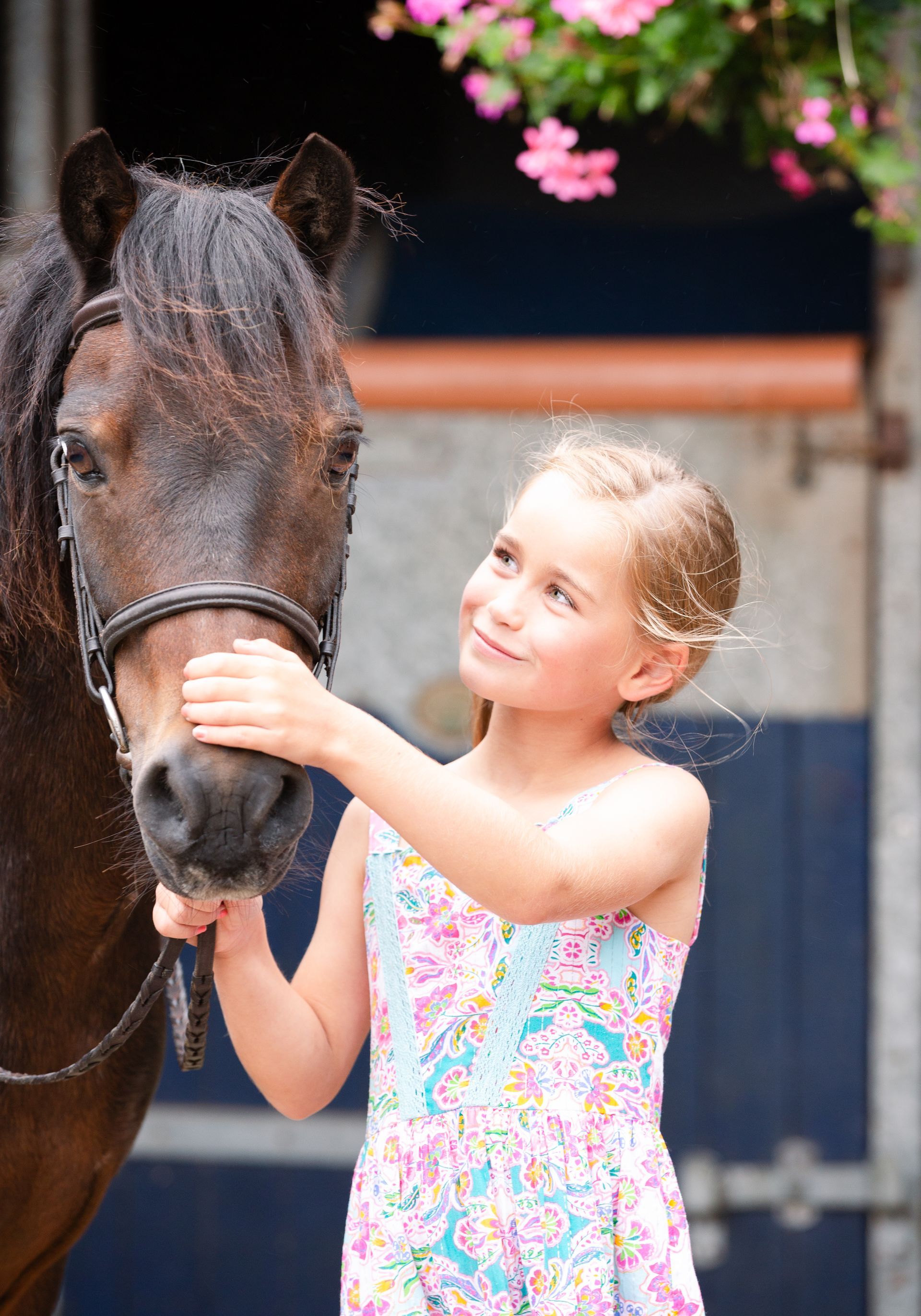 Girl petting a brown pony's nose in a stable, smiling. She wears a floral dress.