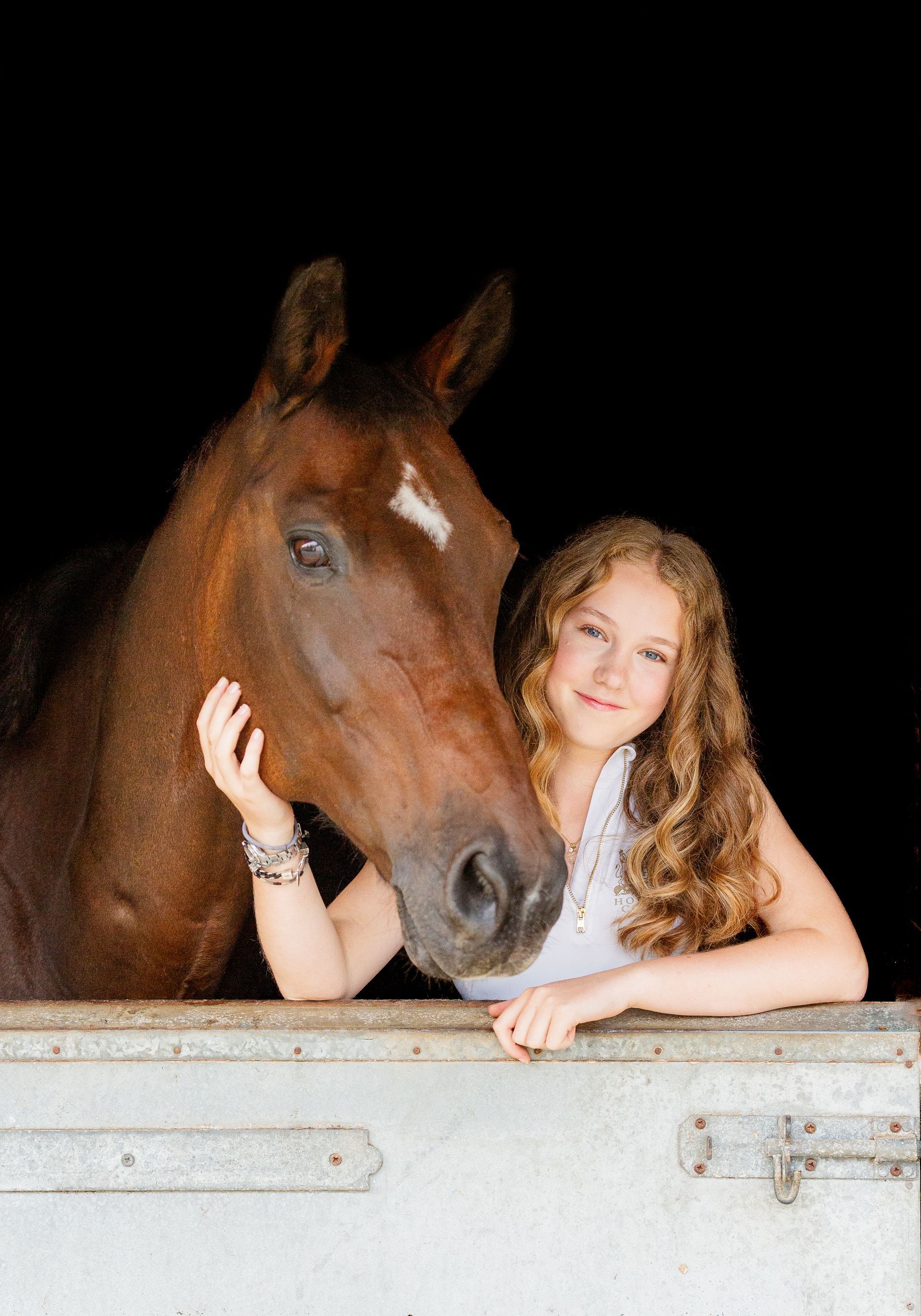 Girl with curly hair smiles, petting a brown horse in a stable.