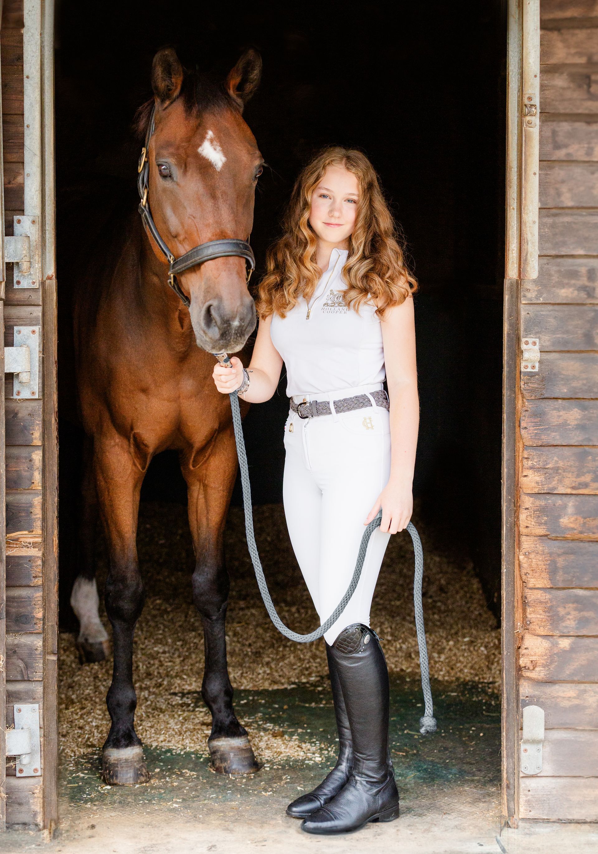 Woman in riding attire with a brown horse in a stable doorway.