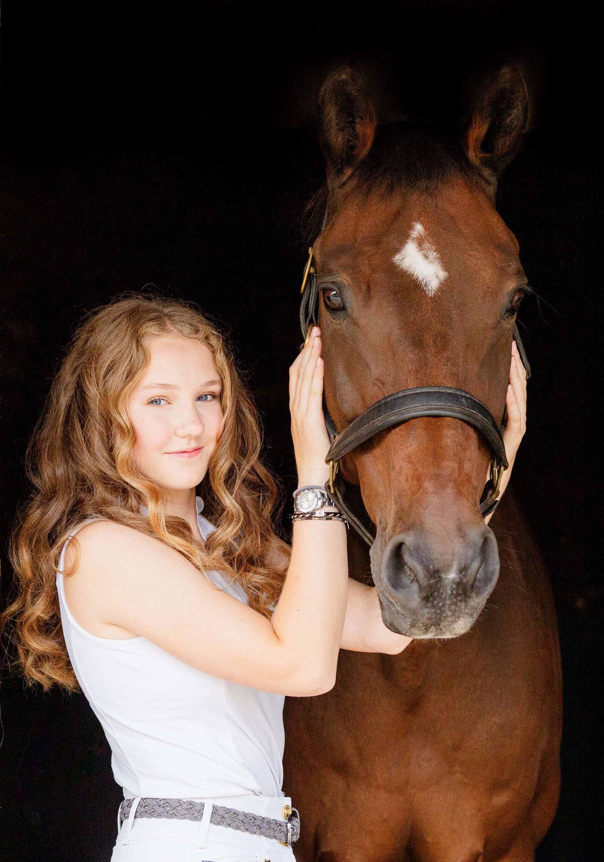 Girl in white top with a brown horse, petting its face in front of a black background.