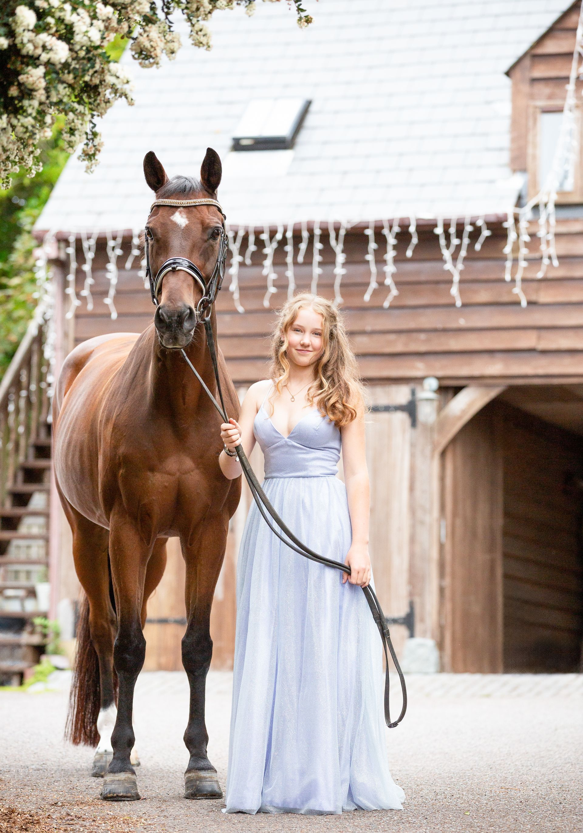 Woman in a blue dress poses with a brown horse in front of a barn.