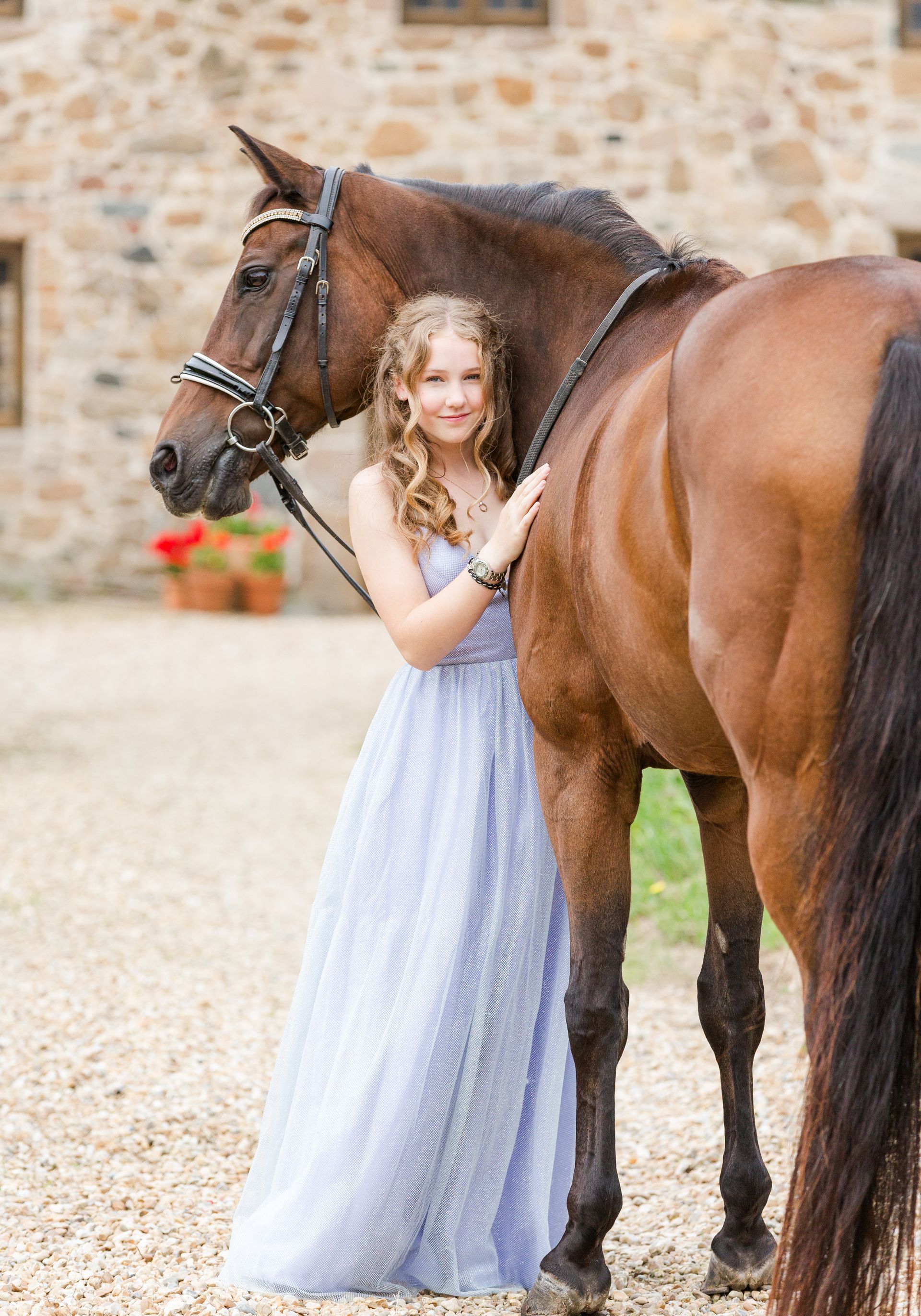 Woman in blue gown with brown horse; standing in front of stone building.
