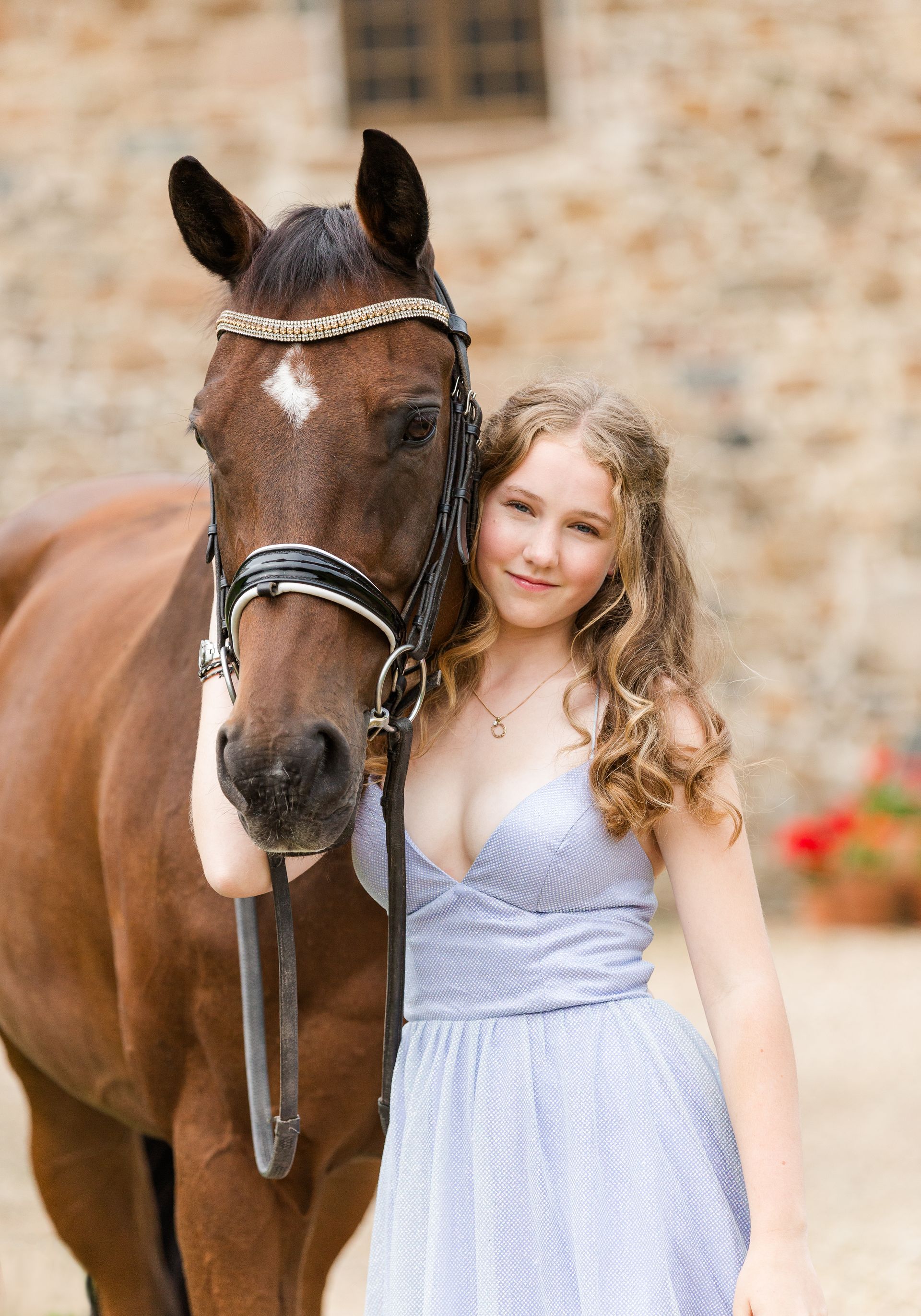 Woman in blue dress stands next to a brown horse, both looking at camera in front of a stone building.