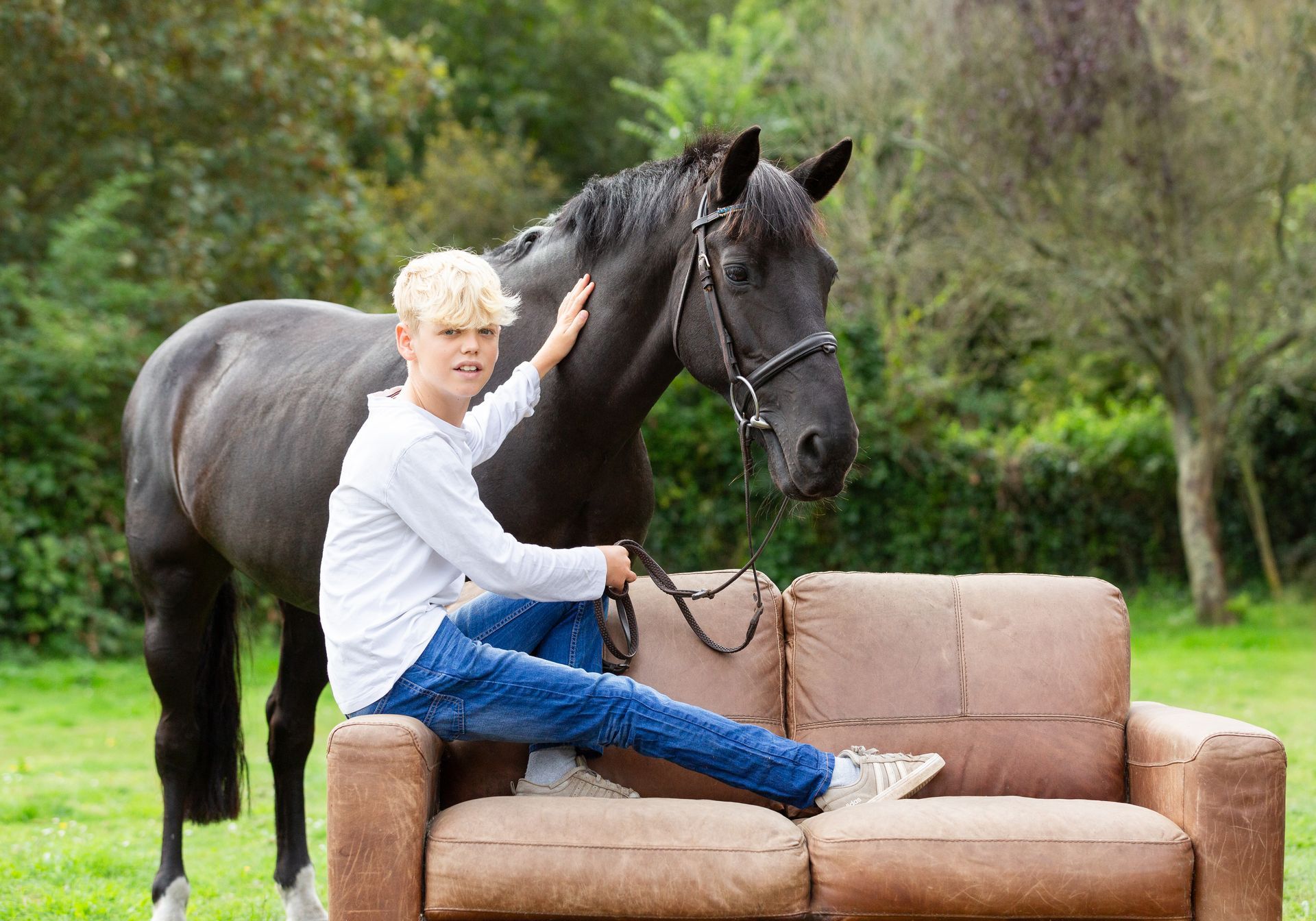 Boy petting a black horse while sitting on a brown sofa outdoors.