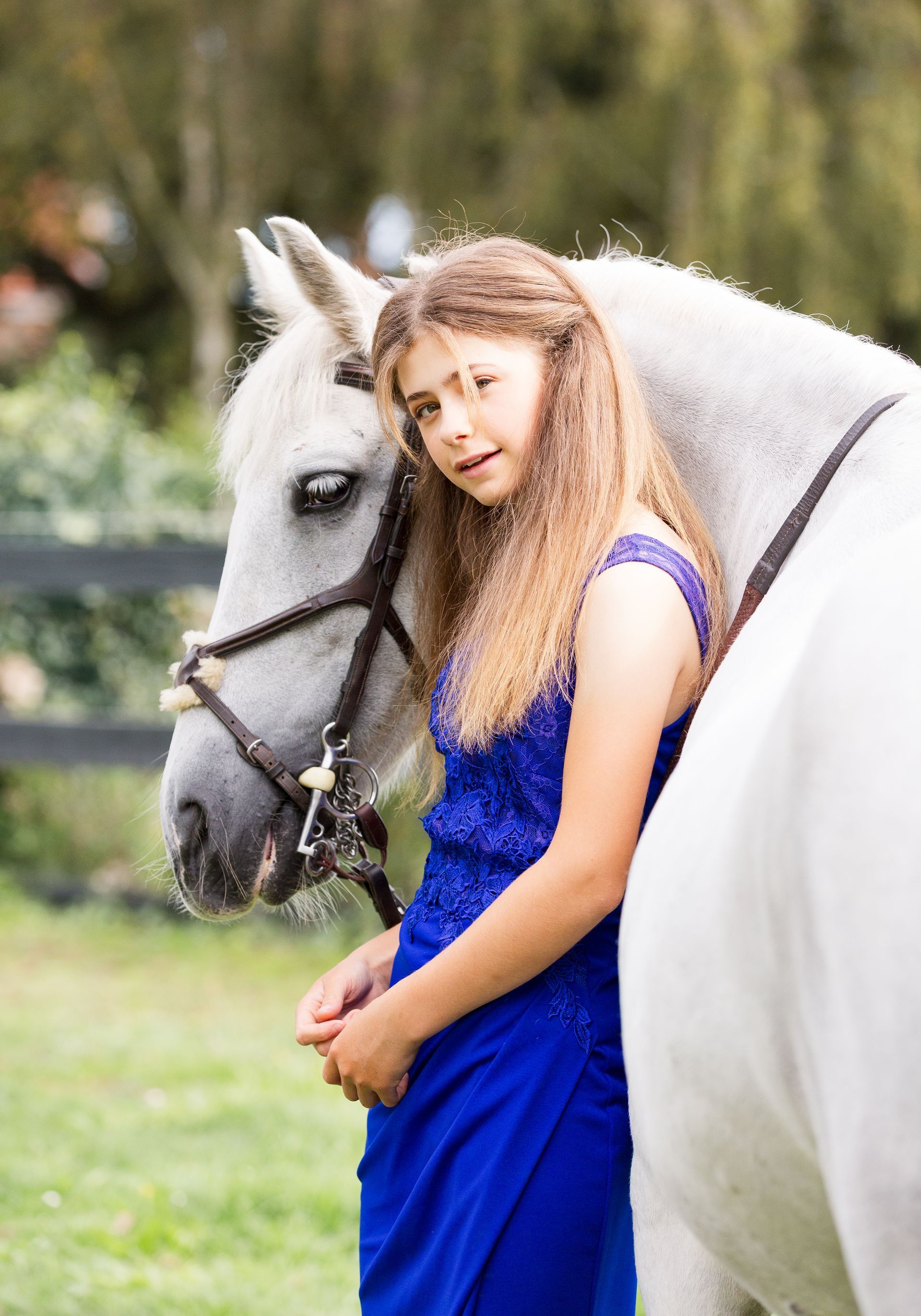 Girl in blue dress leans against a white horse outdoors.