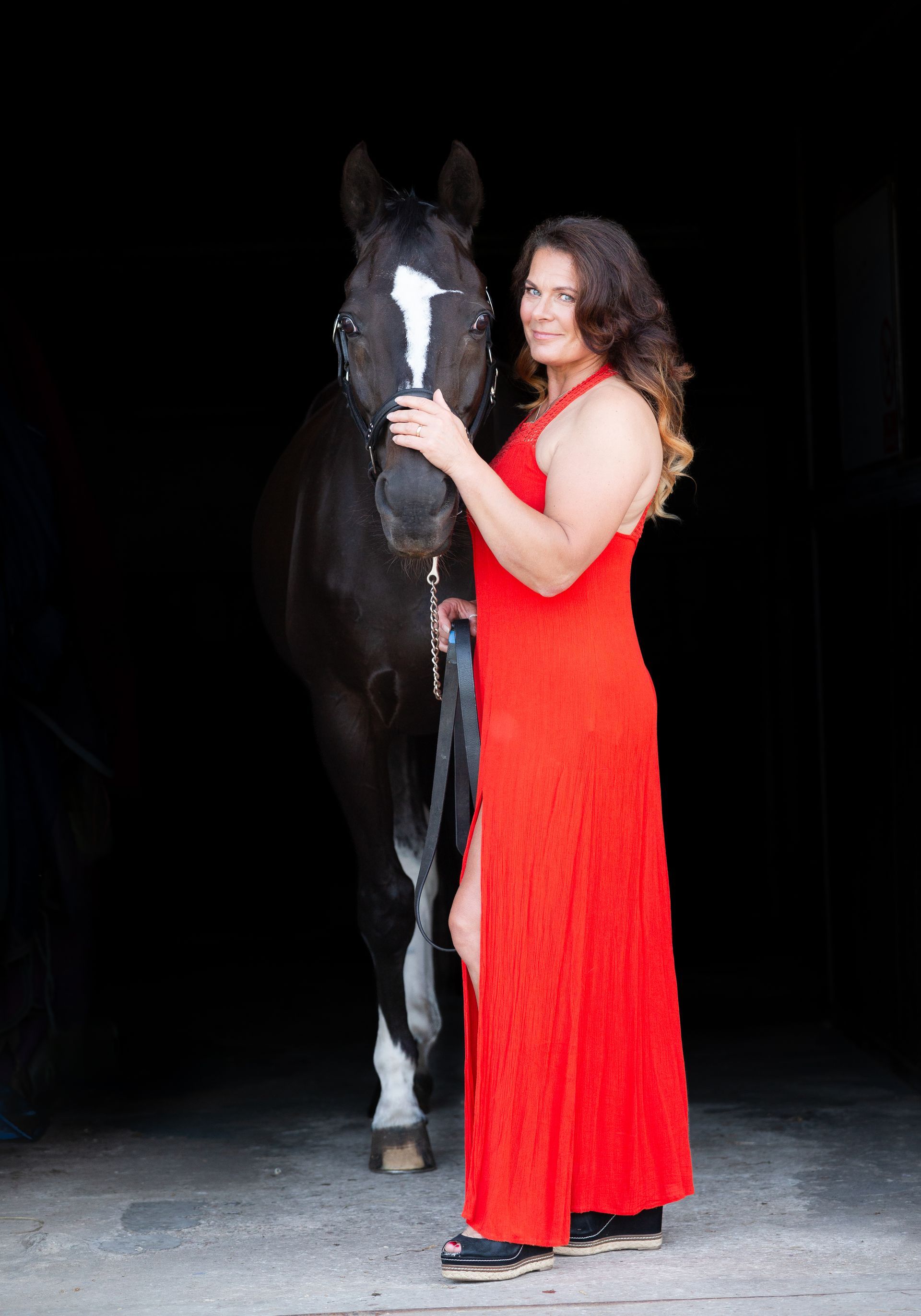 Woman in red dress with black horse in a dark stable.