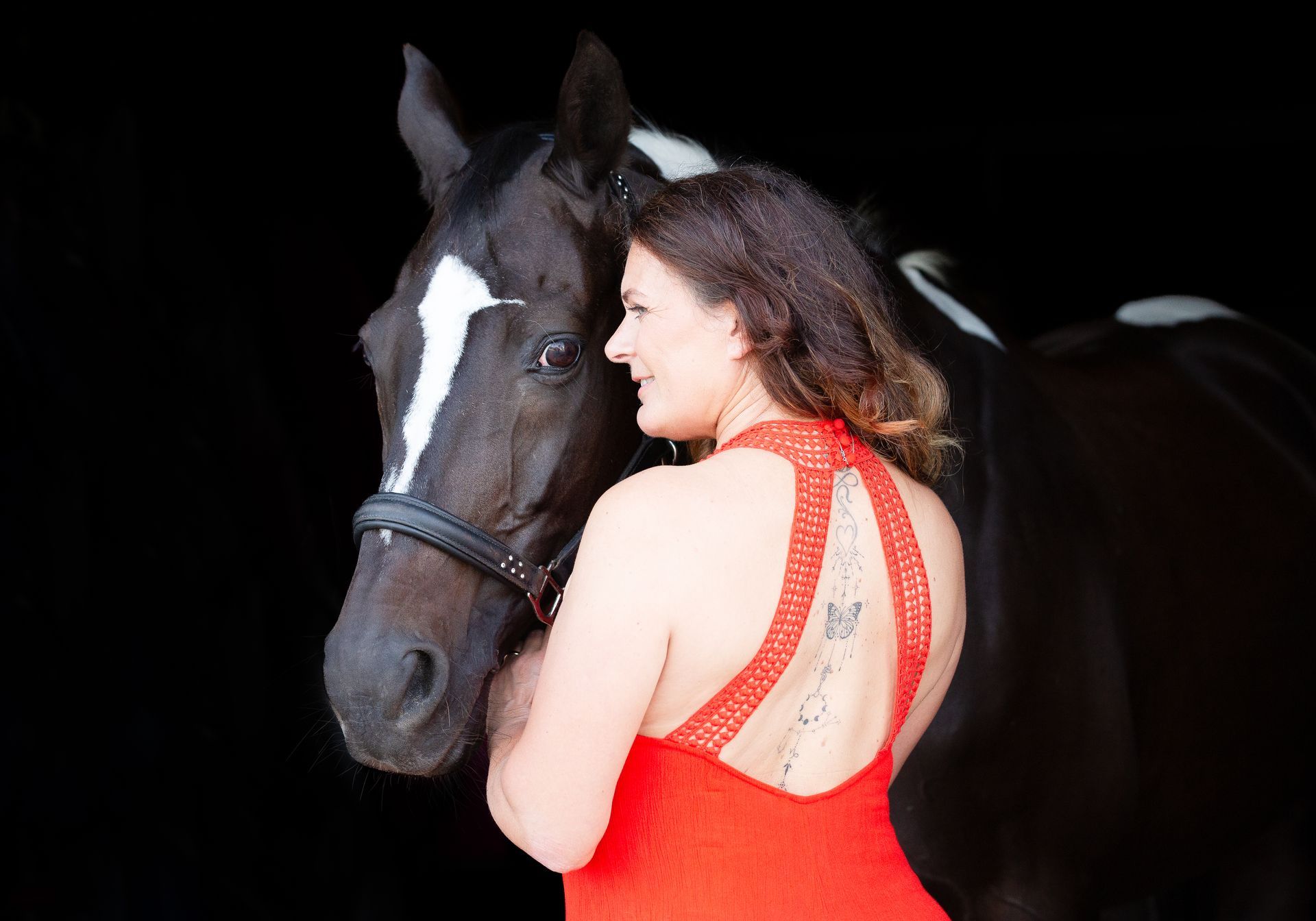 Woman in red dress with black and white horse, smiling at it against black backdrop.