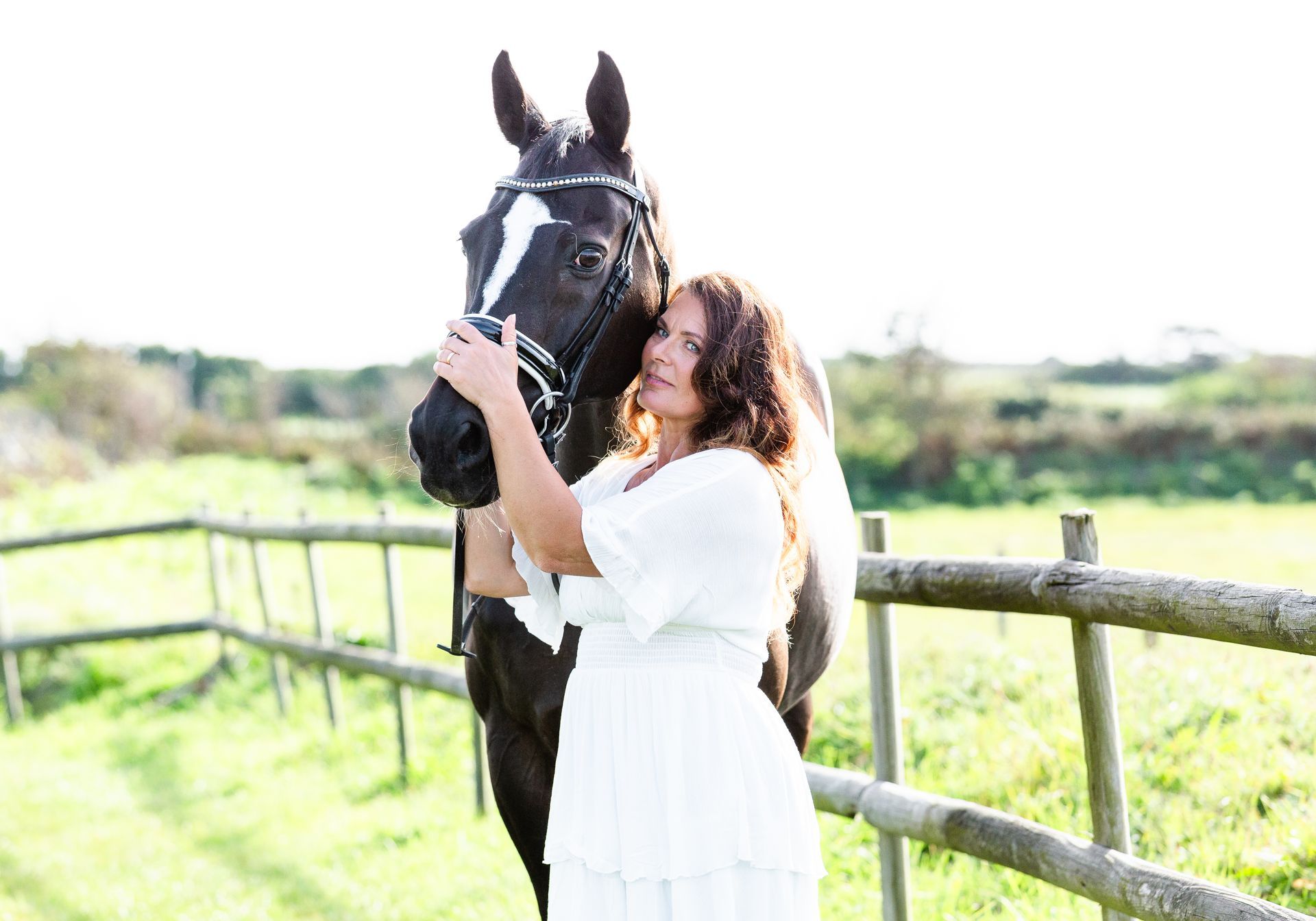 Woman in white dress hugs a dark horse with a white face marking, standing near a wooden fence in a grassy field.