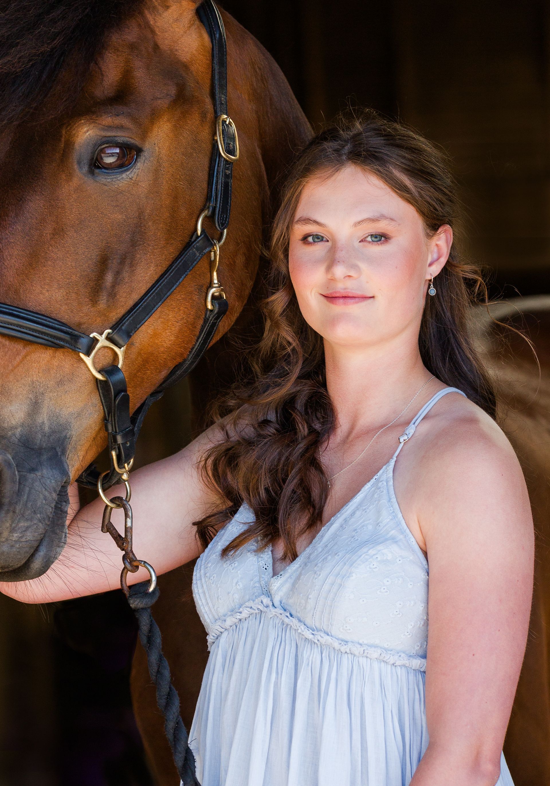 Woman in a light blue dress poses with a brown horse in a barn.