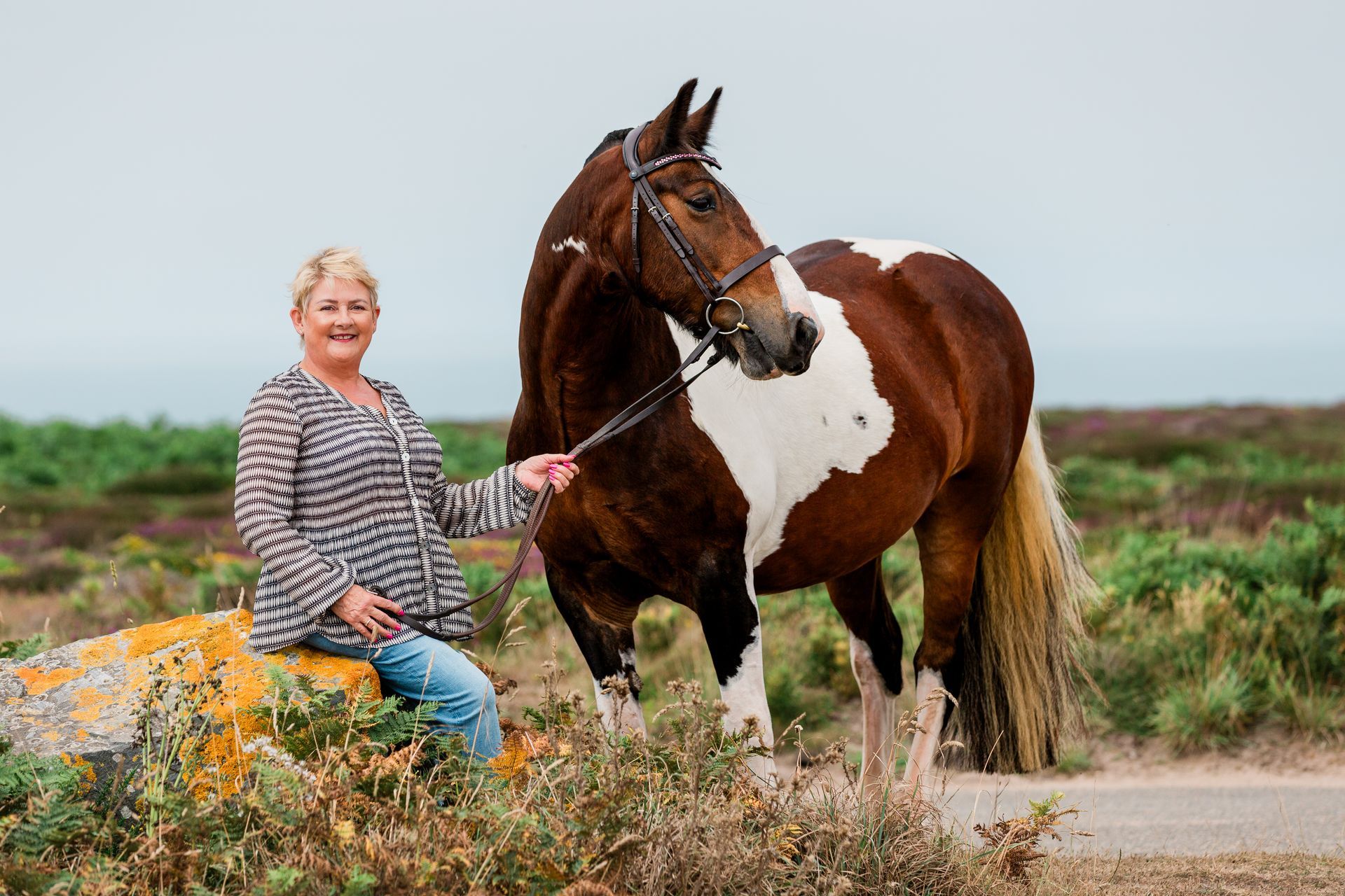 Woman sits by a colorful pinto horse outdoors, holding the lead. Coastal landscape background.