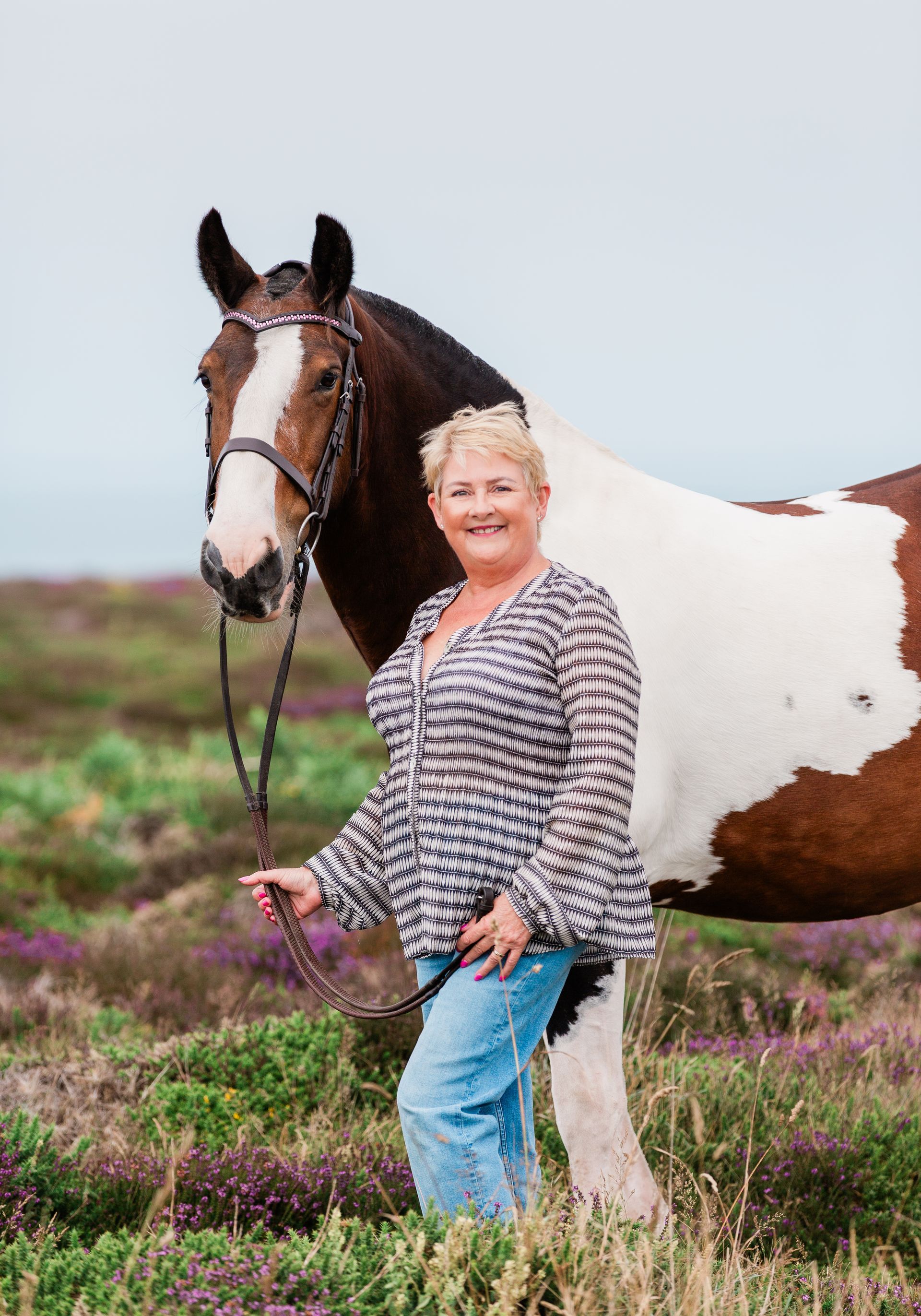 Woman in patterned shirt and jeans holding a horse's reins in a field of purple flowers.