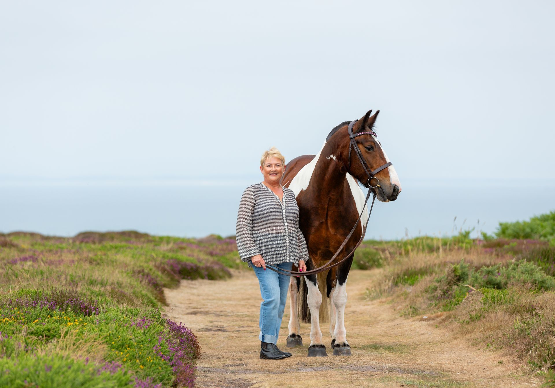 Woman stands beside a piebald horse on a path with ocean backdrop.