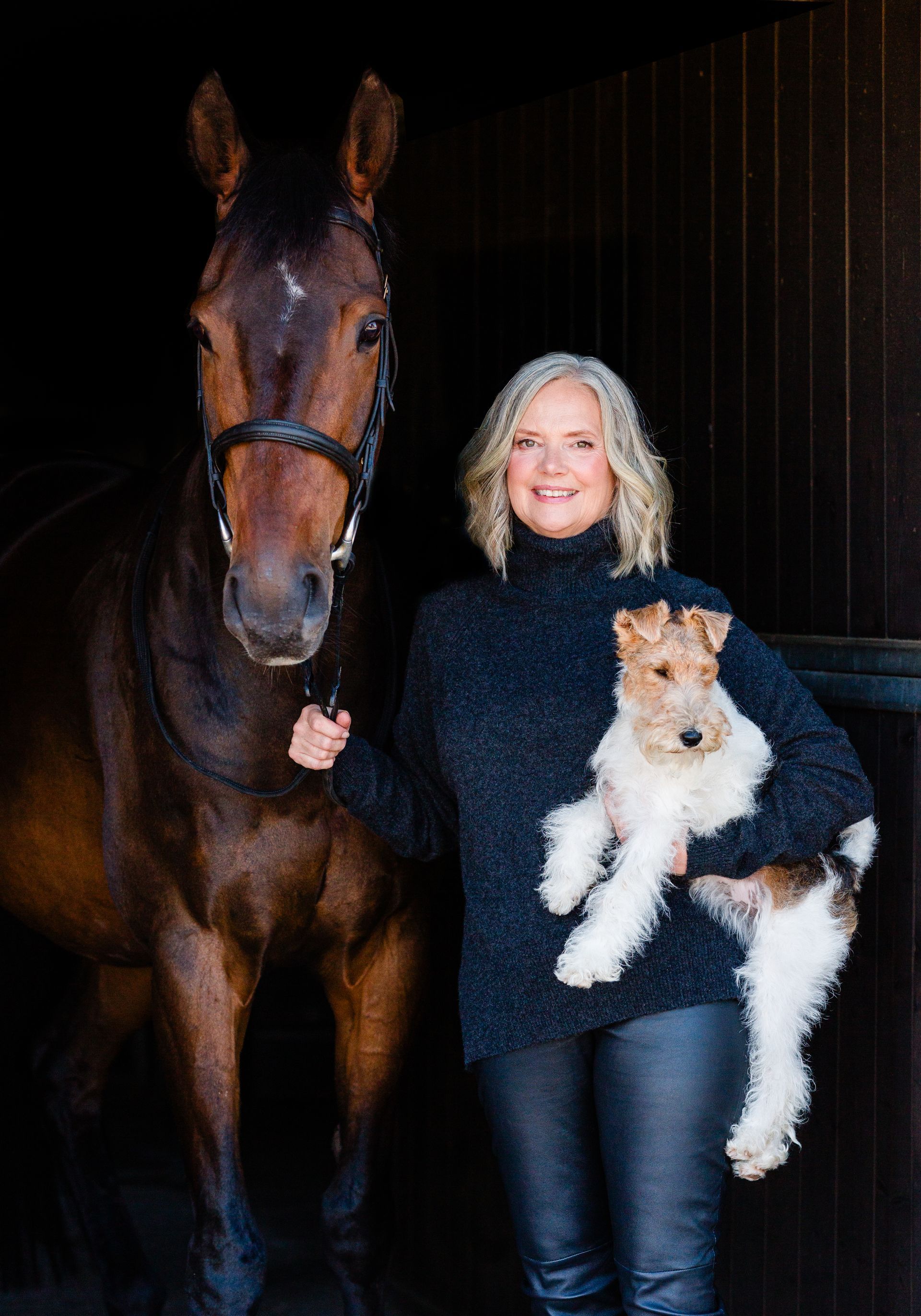 Woman in dark sweater holding a small dog, with a horse in a stable.