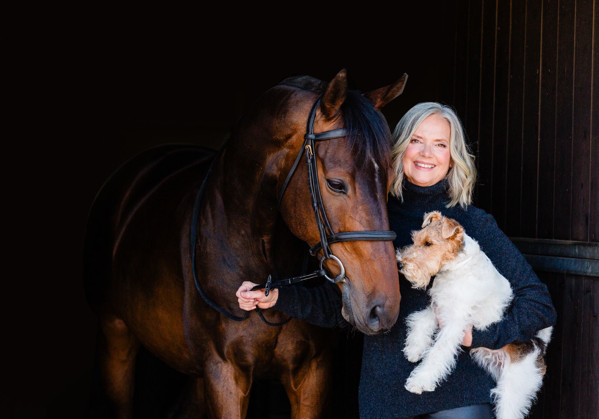 Woman with a horse and small dog in a stable; woman smiles, holds dog; horse has bridle; dark background.