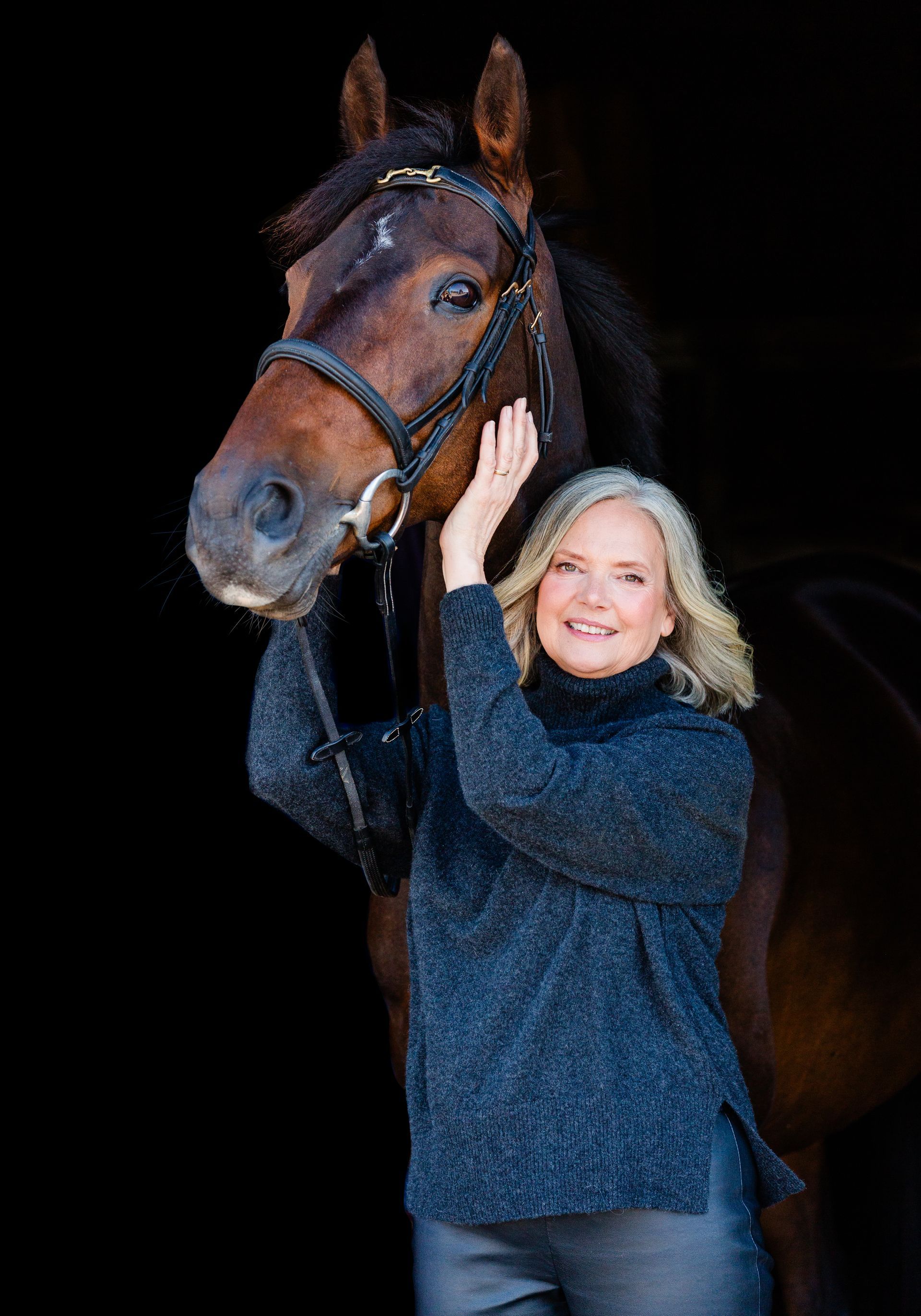 Woman in blue sweater petting a brown horse in front of a black background.