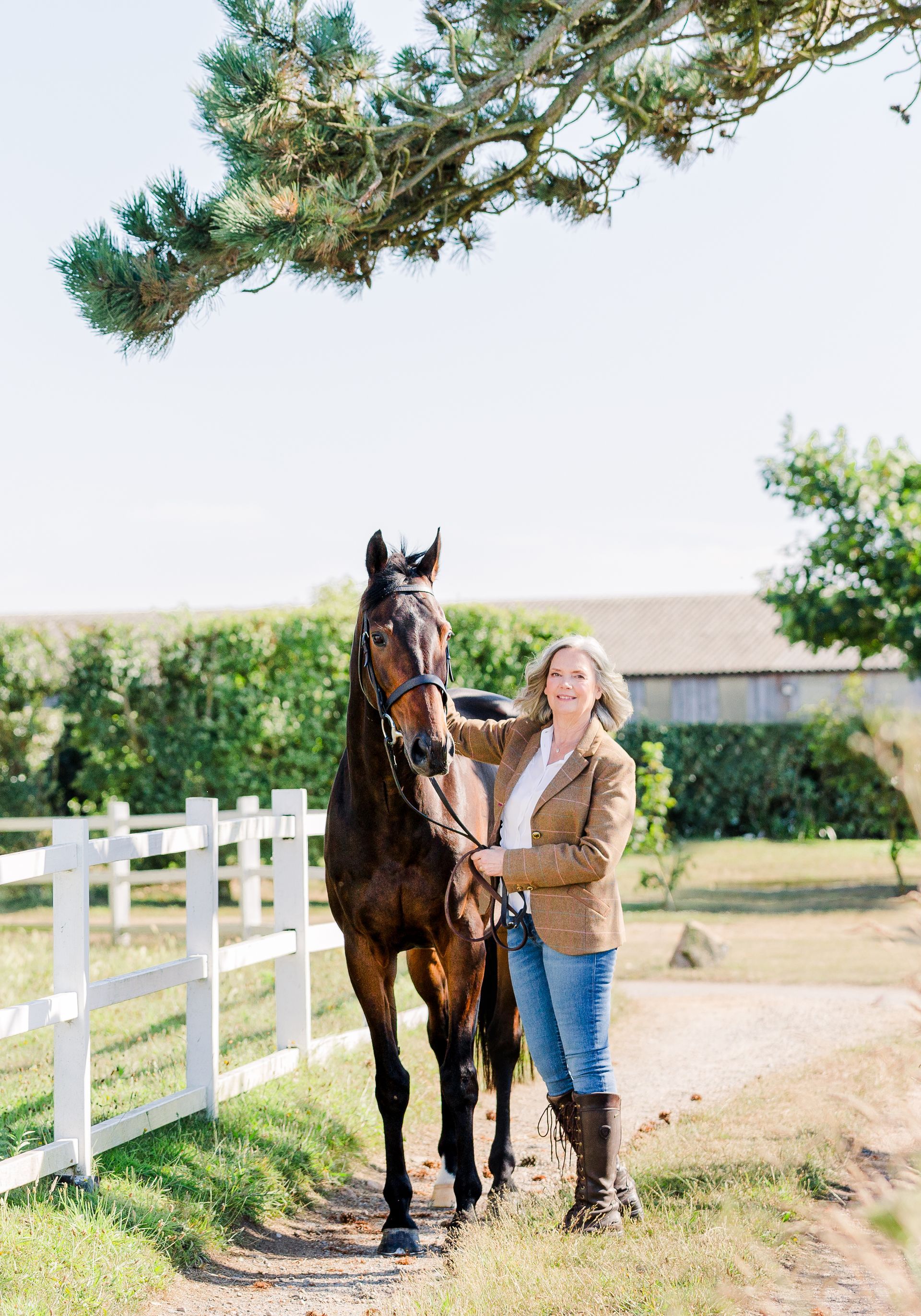 Woman in jacket and jeans stands next to a dark horse near a white fence, under a tree.