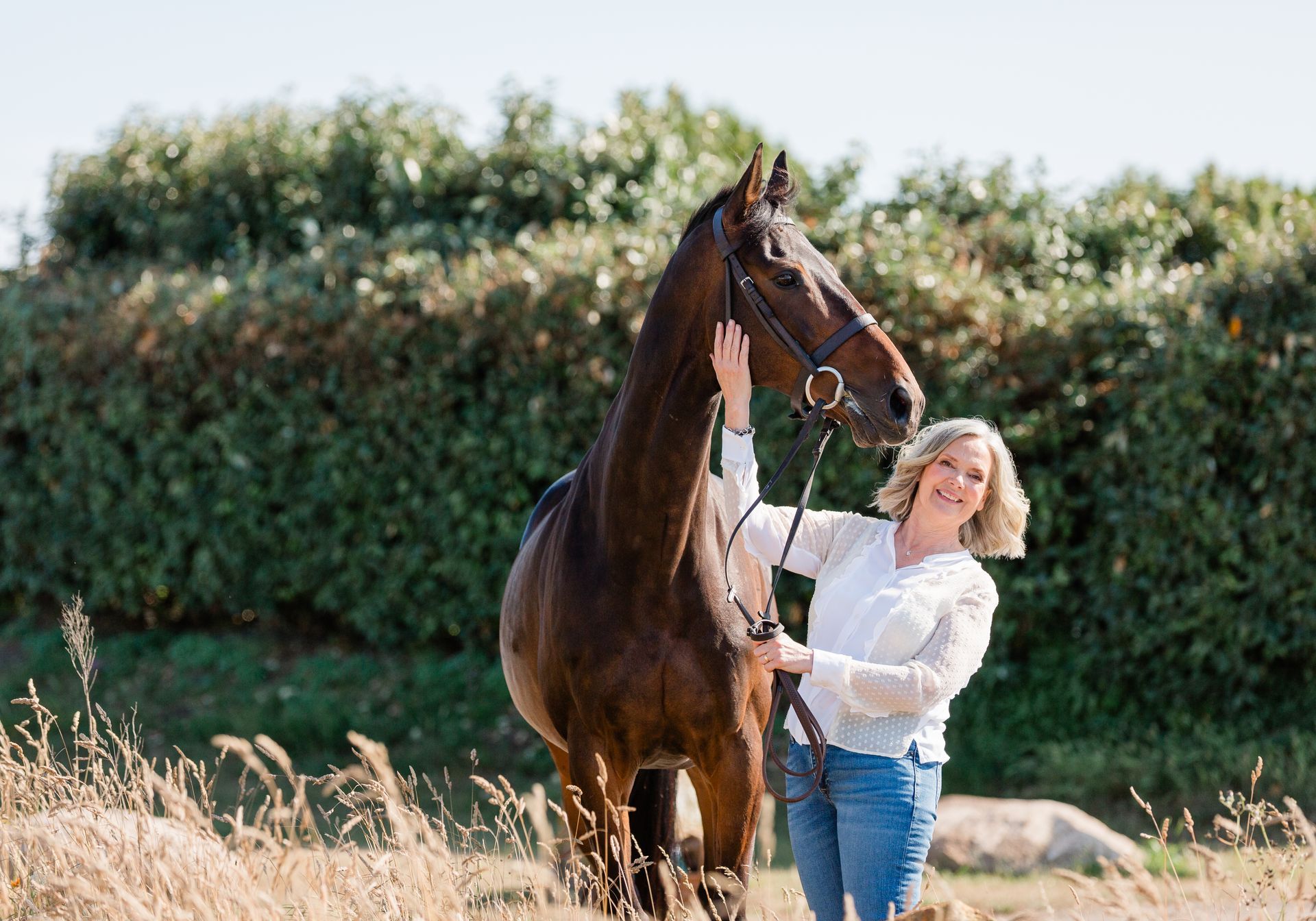 Woman with blonde hair smiles, holding reins of a brown horse in a field with green foliage.