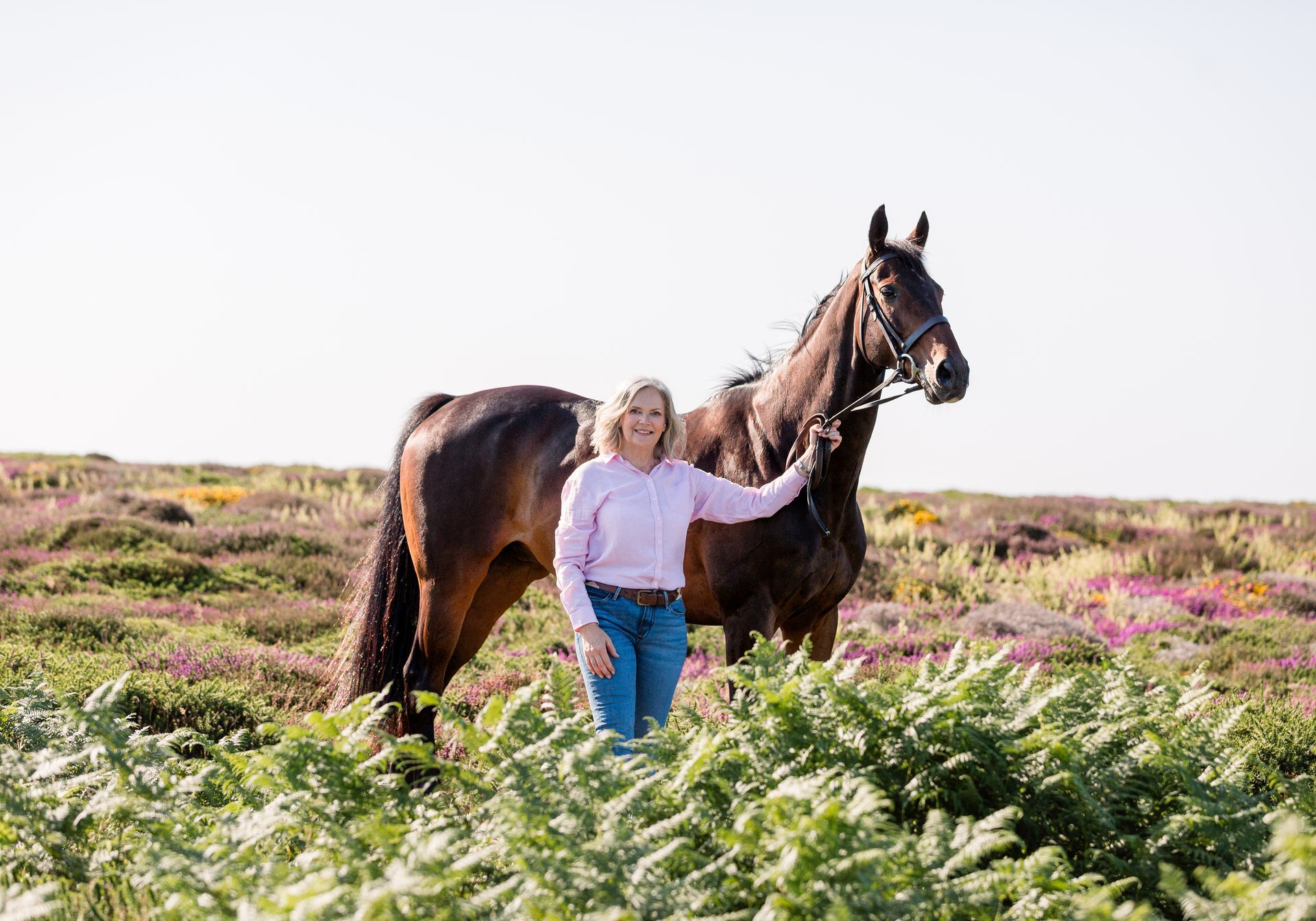 Woman in pink shirt and jeans poses with a brown horse in a field of purple flowers.