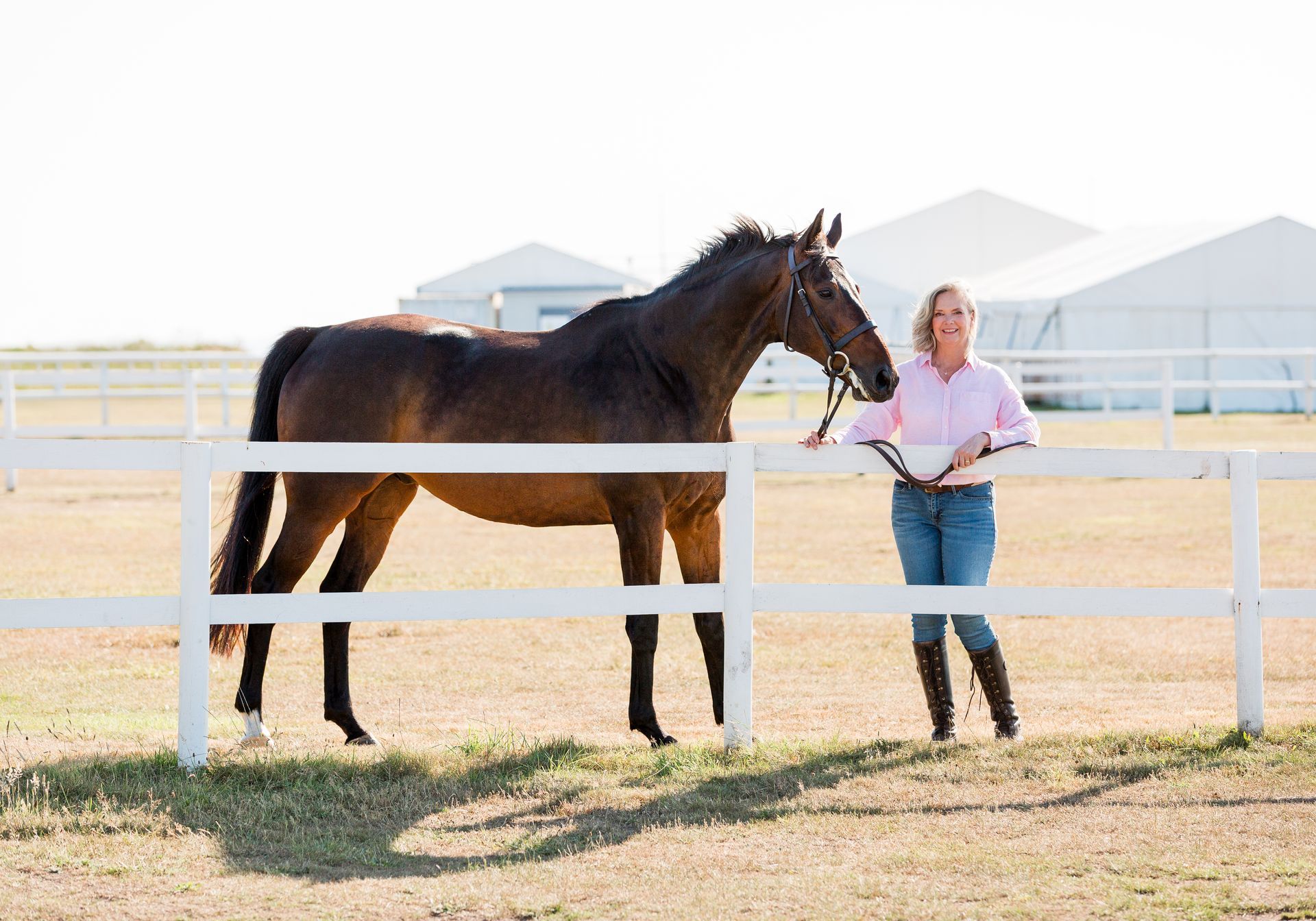 Woman in pink shirt and jeans holding a bridle next to a brown horse behind a white fence in a field.