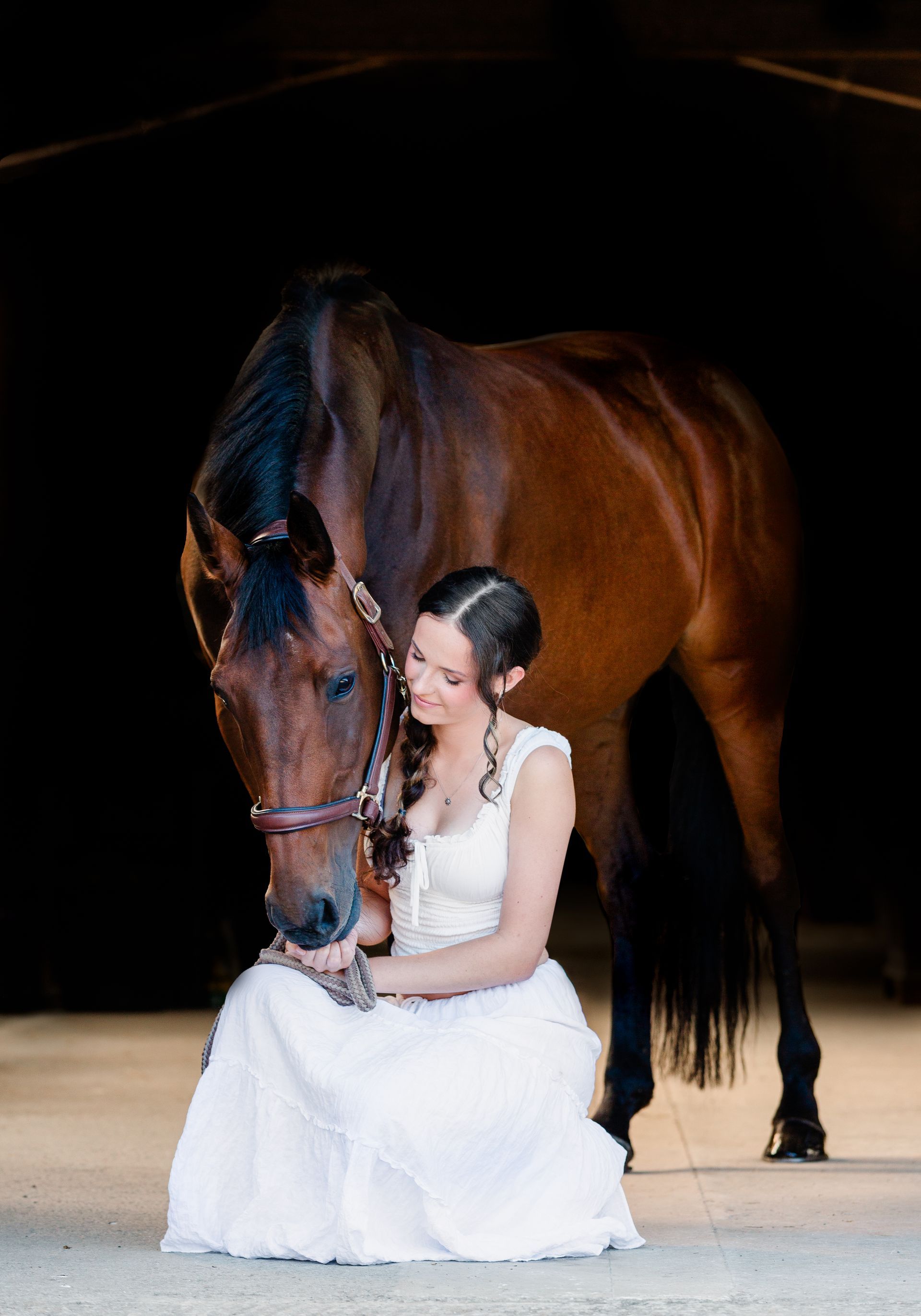 Woman in a white dress kneels, petting a brown horse; dark background.