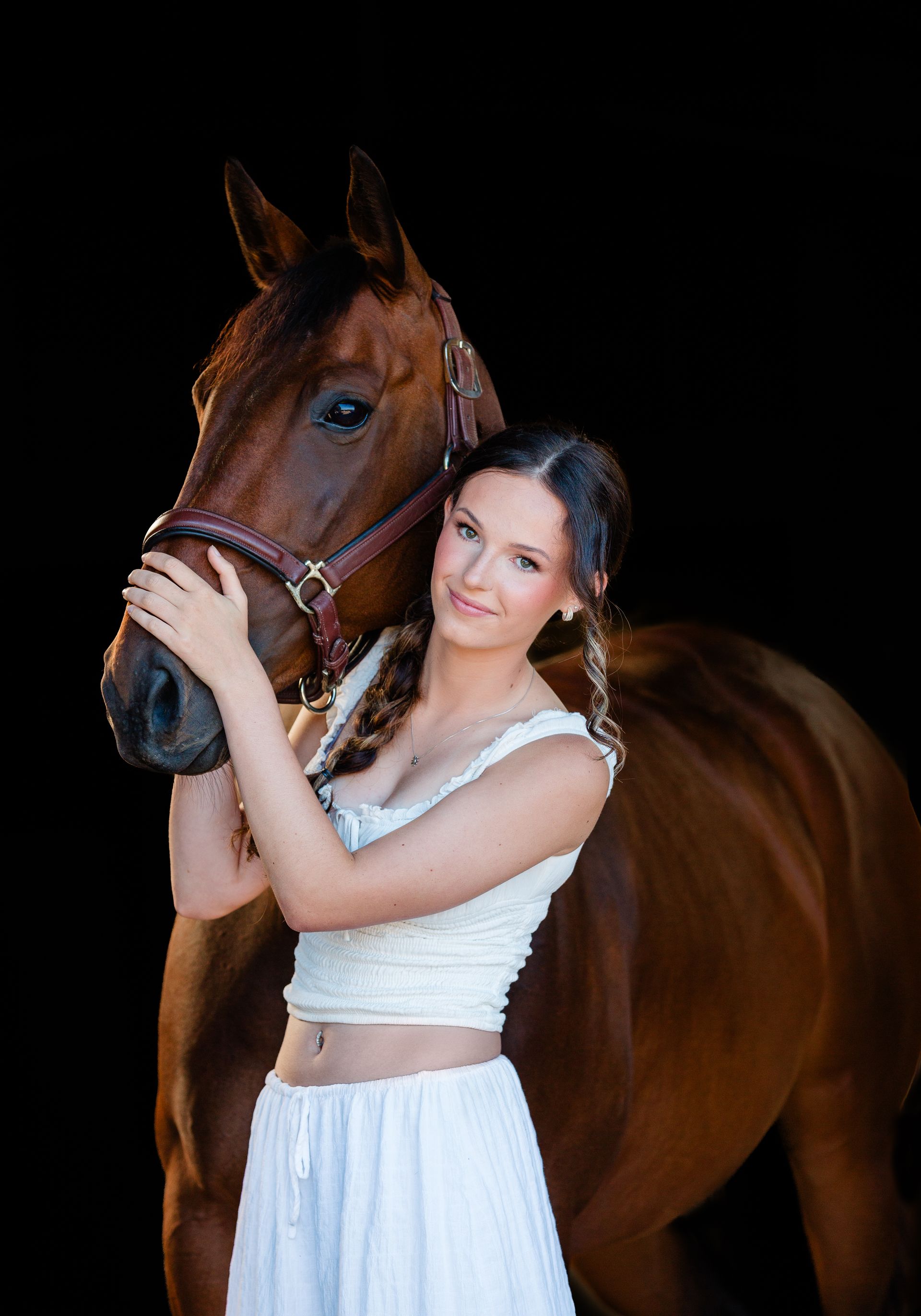 Woman in white outfit with brown horse, embracing and smiling against a black backdrop.