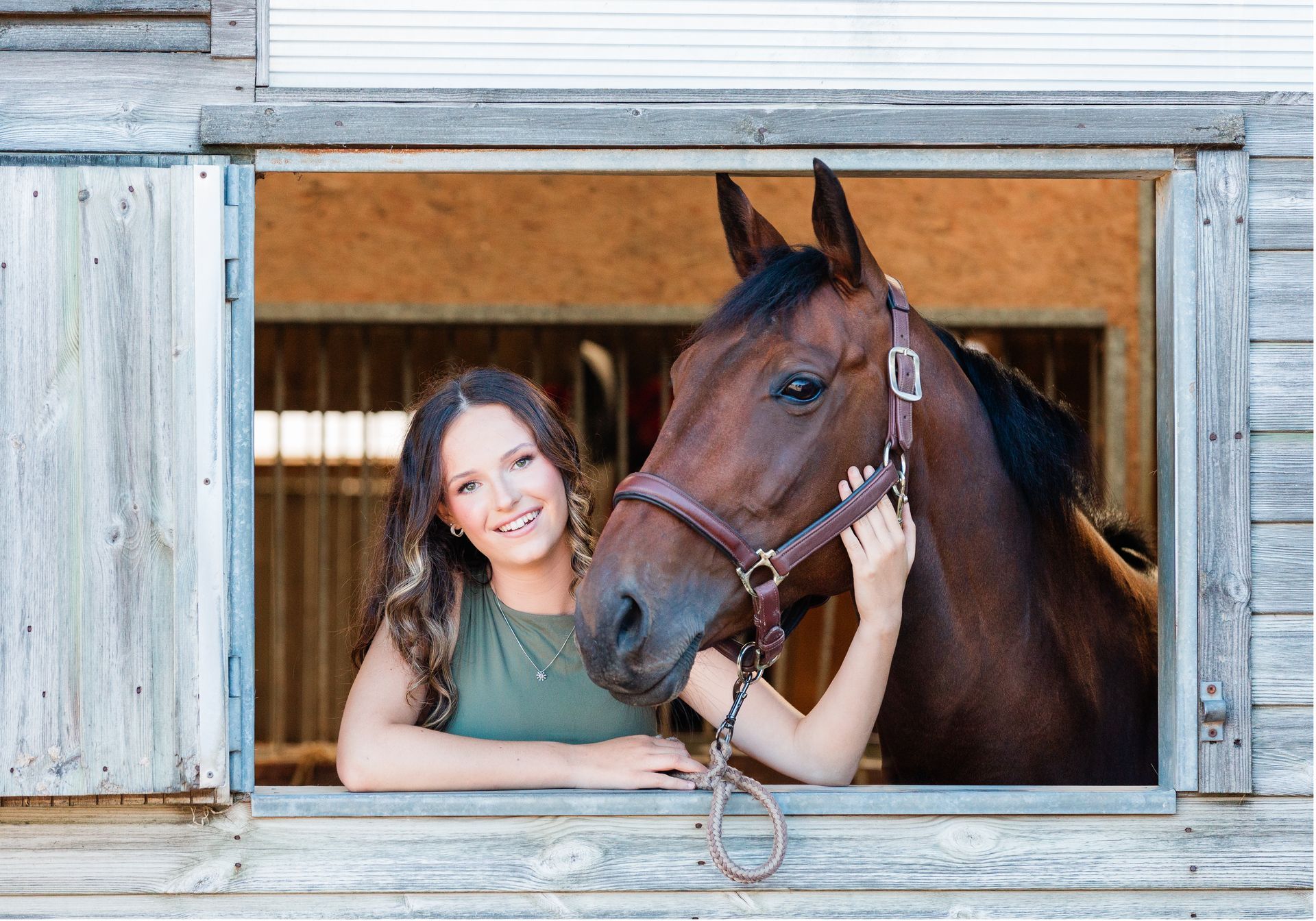 Woman in green top smiles, pets brown horse's head; both at stable window.