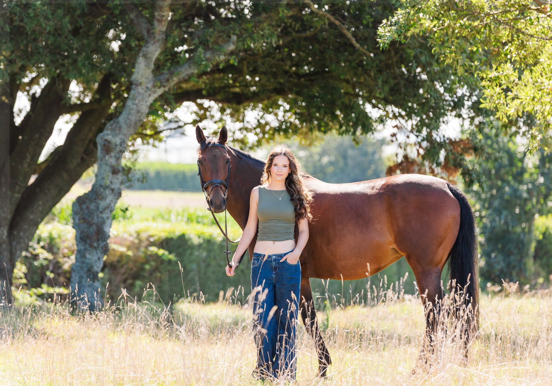 Woman in jeans and green top stands beside a brown horse under a tree in a field.