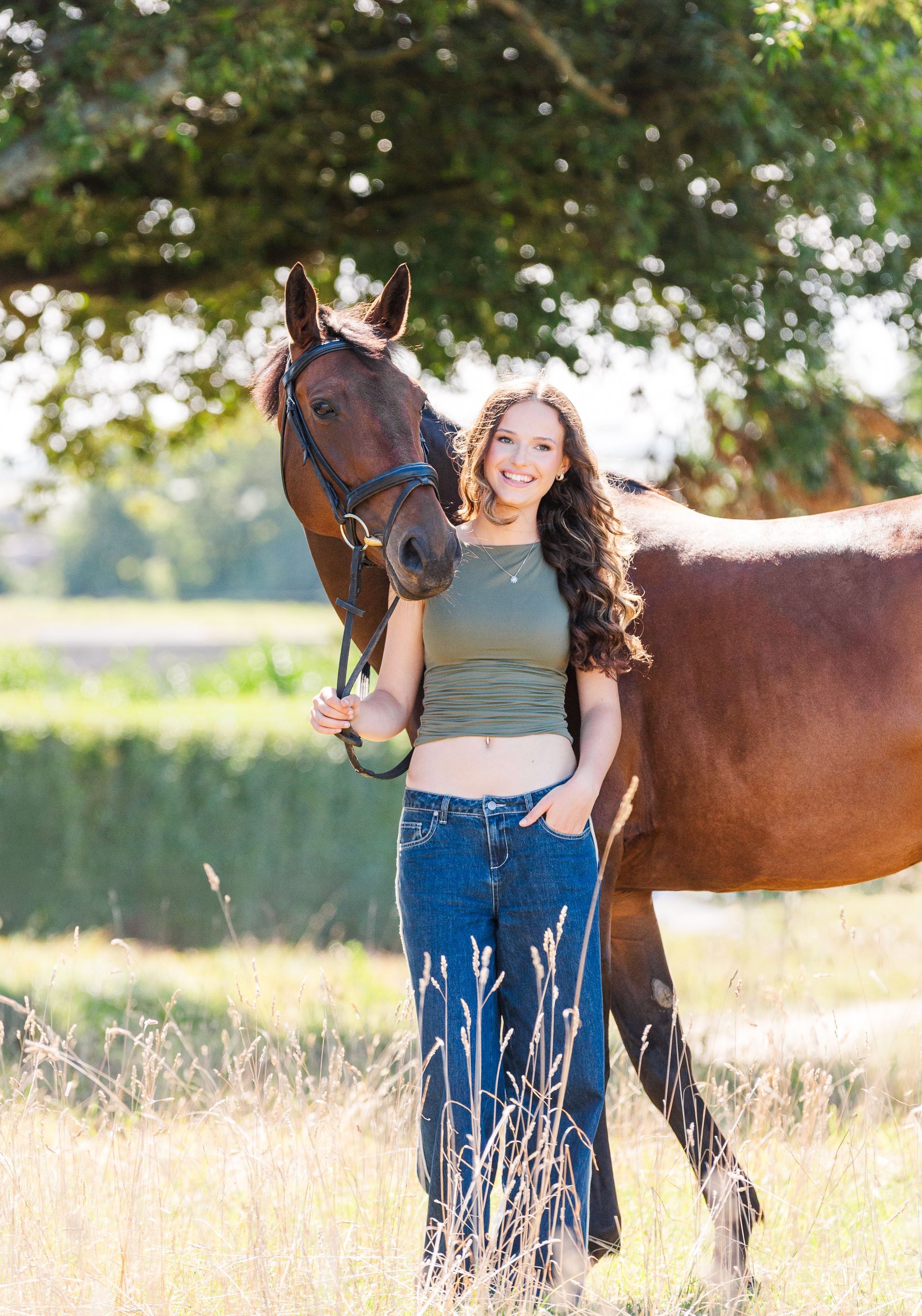 Woman with curly hair and horse in field. She wears a green top and jeans. Sunlit, outdoors.
