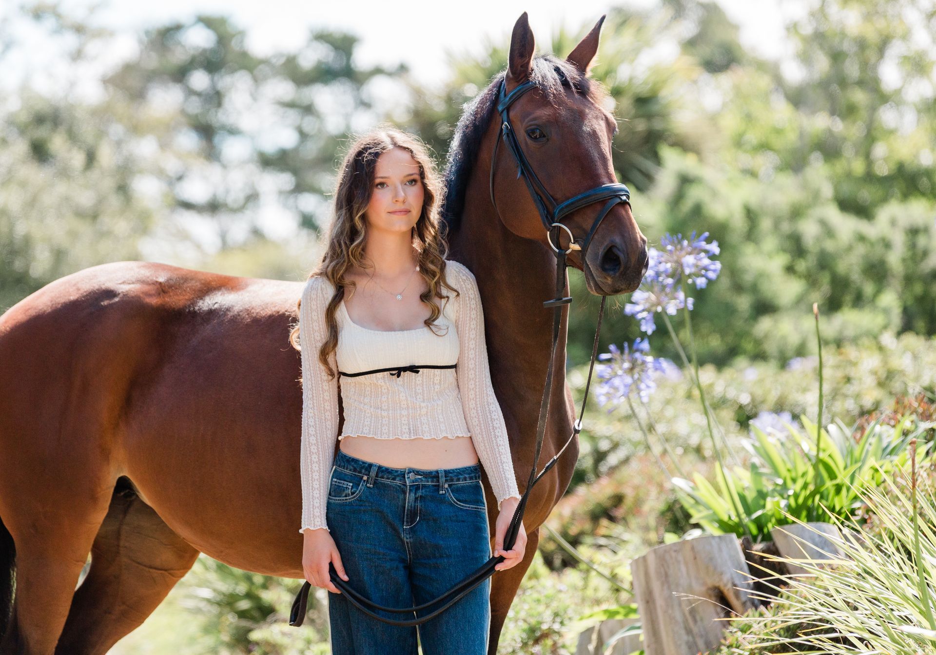 Woman in jeans and lace top stands next to a brown horse in an outdoor setting.
