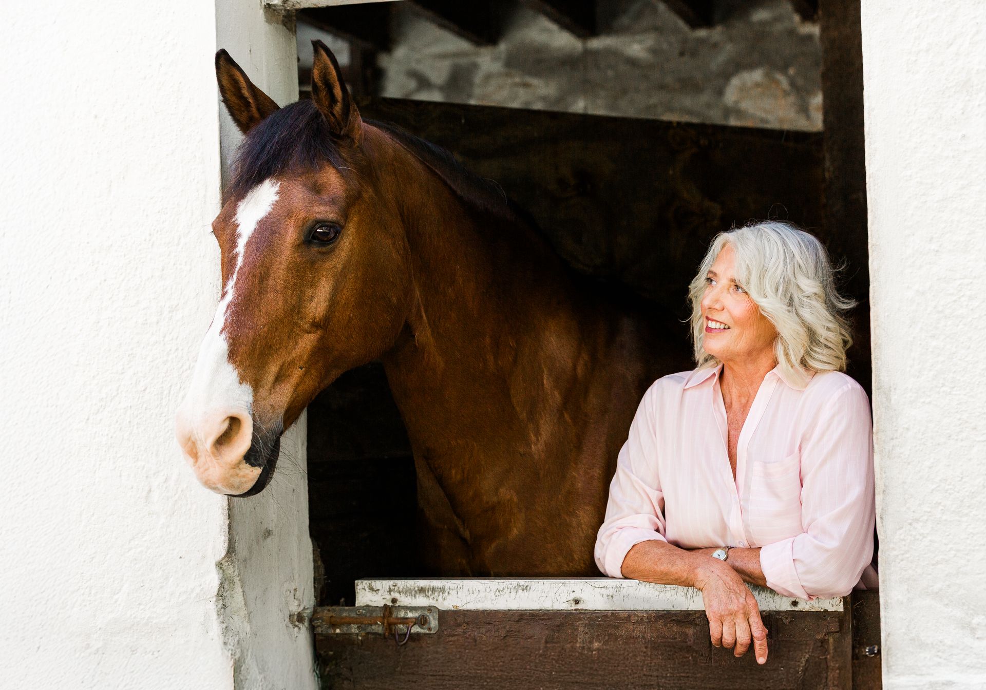 Woman in a pink shirt leaning on a stable window beside a brown horse with a white blaze.