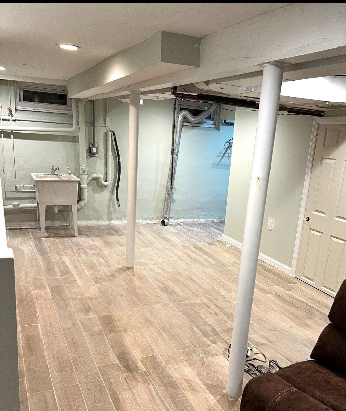 Basement with light wood floor, support beams, utility sink, open doorway, and a recliner.