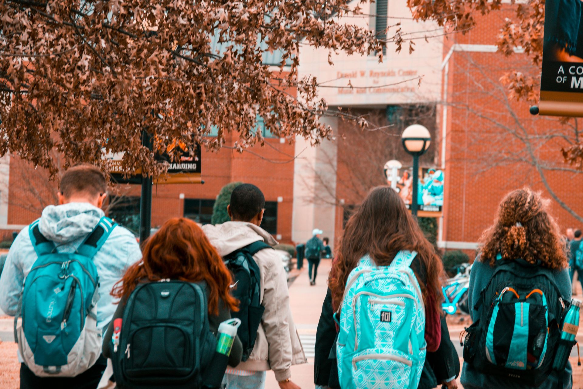 Students walking with backpacks toward a brick building on a college campus with fall foliage.