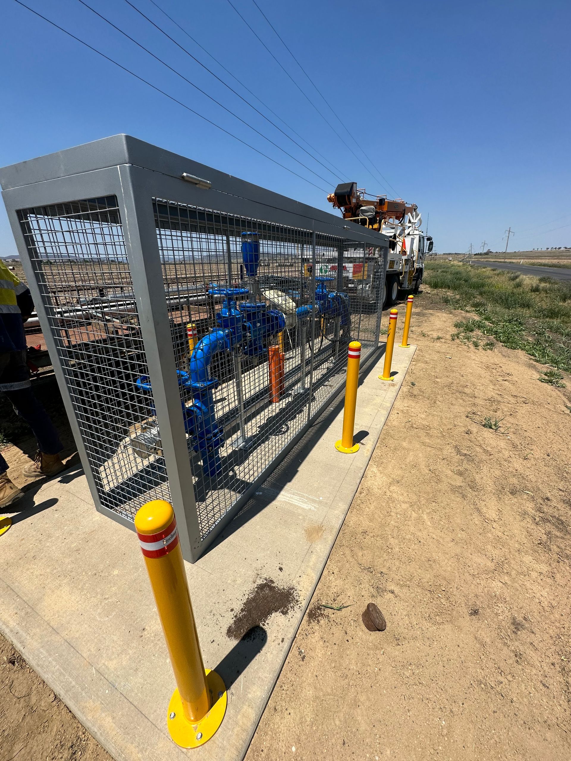 Blue industrial equipment in a fenced utility enclosure beside a dirt path and yellow bollards.