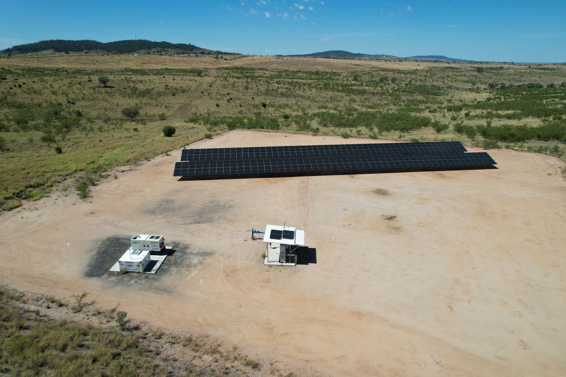 Solar panels and two small structures on a flat, sandy field under a clear sky