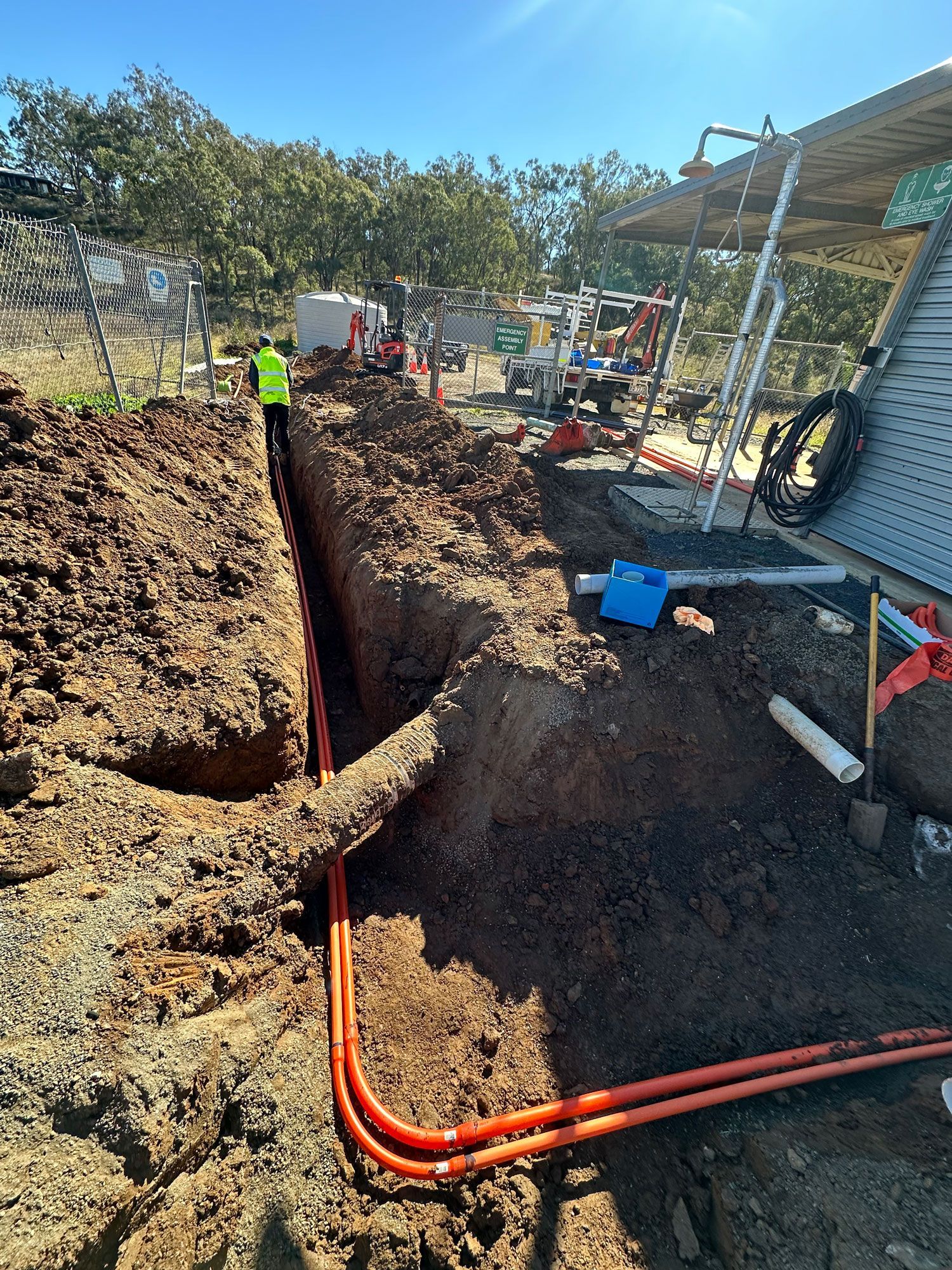 Workers trenching beside a house, with orange conduit pipes and excavated dirt under bright sunlight