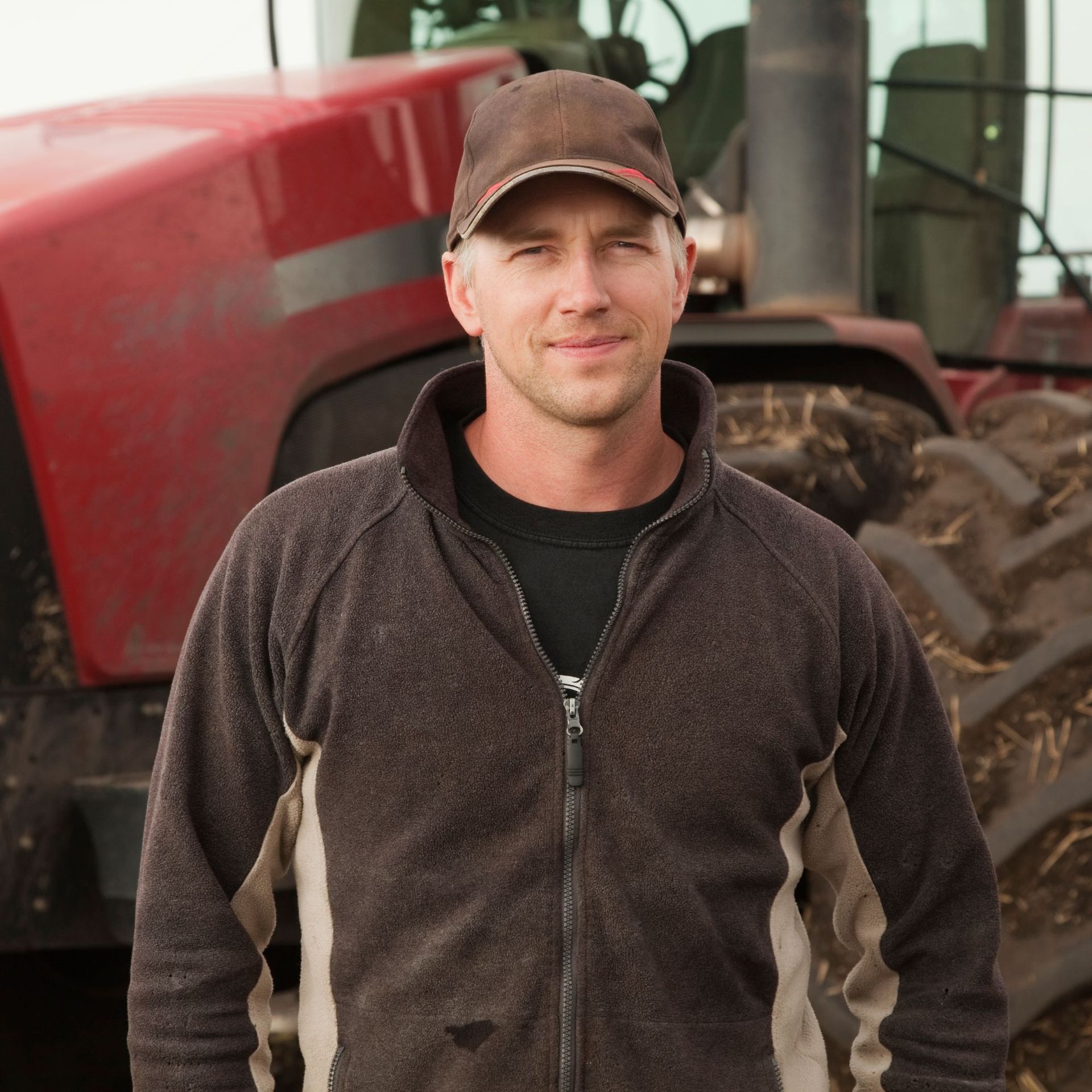 Man in cap and zip-up jacket smiles in front of a red tractor.