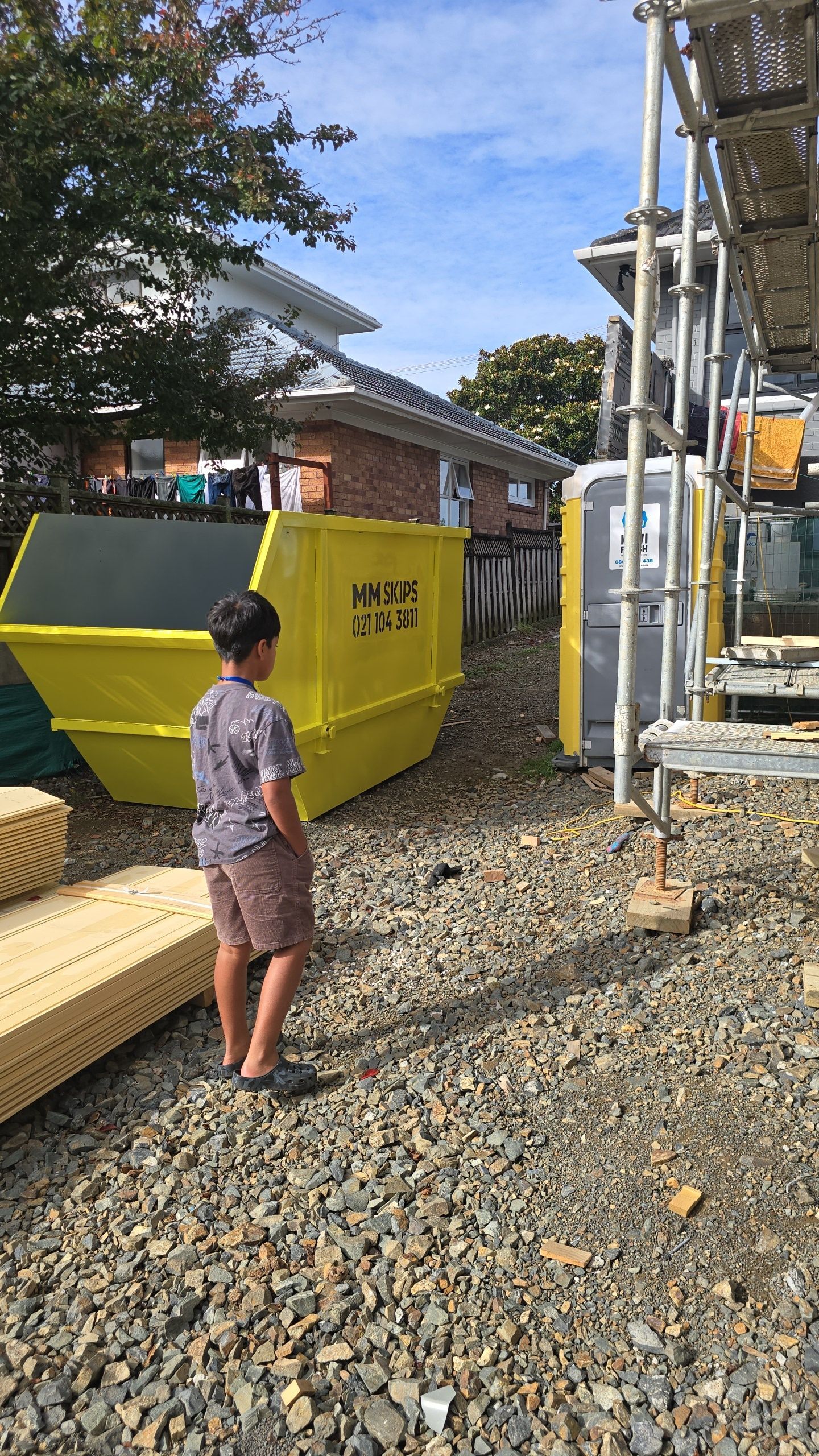 Person stands on gravel near a yellow bin and metal scaffolding by a small building.