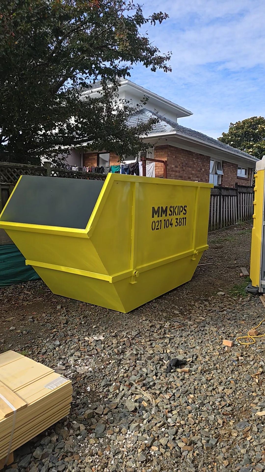 Yellow construction dumpster beside a house, with gravel ground and trees in the background