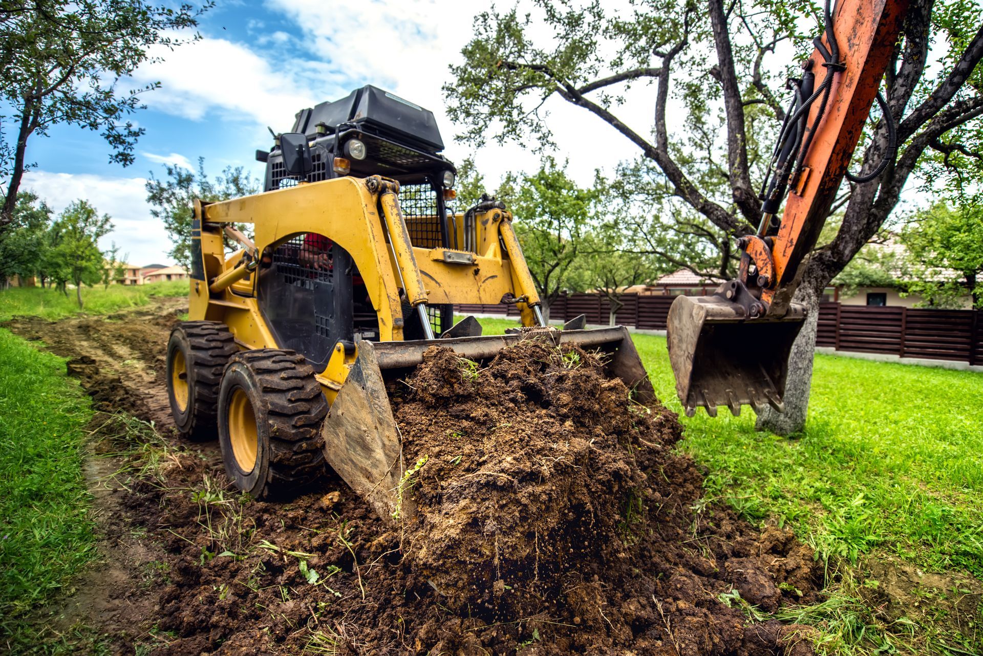 Yellow excavator digging dirt in a grassy yard with trees and a black dump truck nearby
