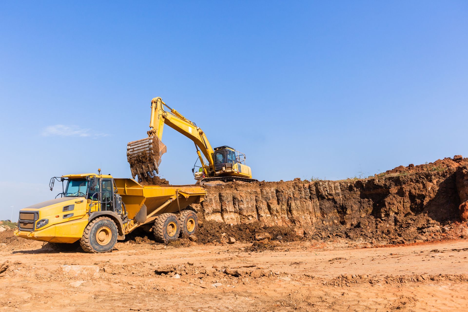 Yellow excavator and dump truck working on a dirt construction site under a blue sky