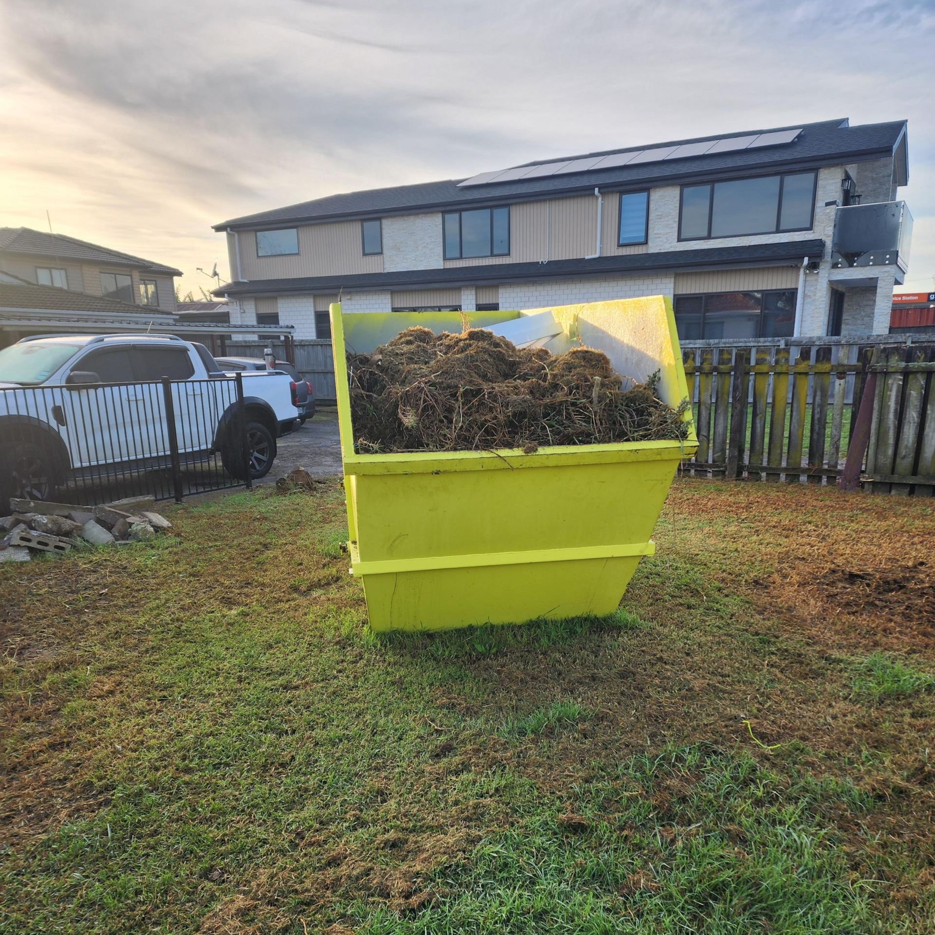 Yellow dumpster filled with yard debris on a grassy lot in front of row houses at sunset