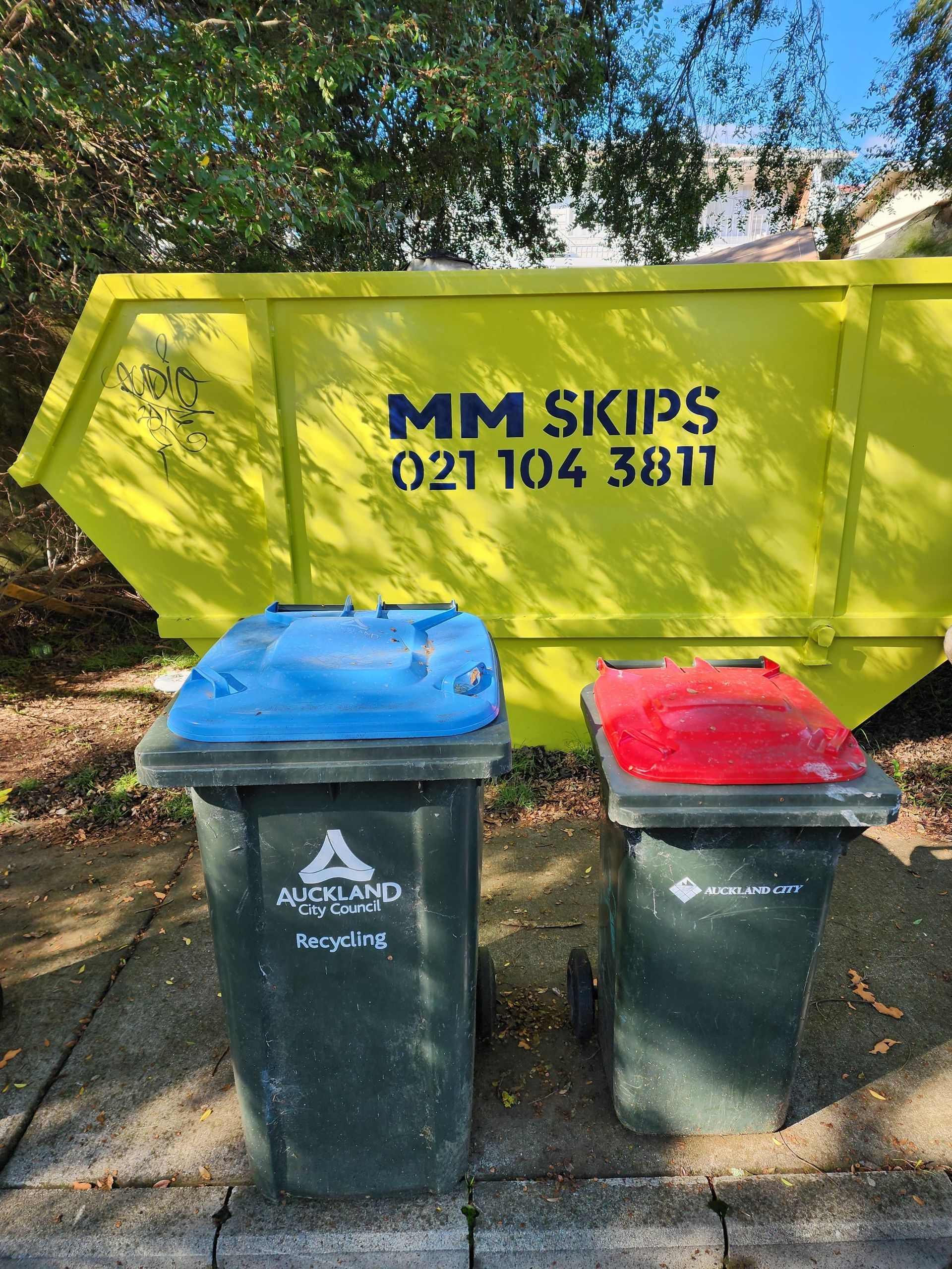Two outdoor rubbish bins with blue and red lids beside a yellow MM Skips dumpster.