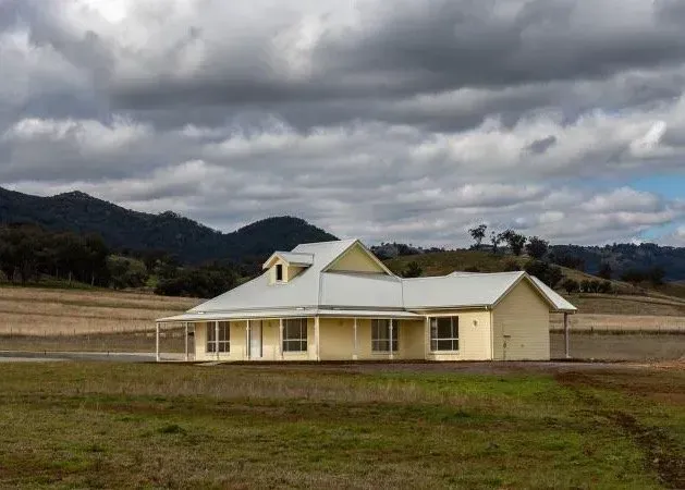 A yellow house with a white roof is in the middle of a grassy field with mountains in the background