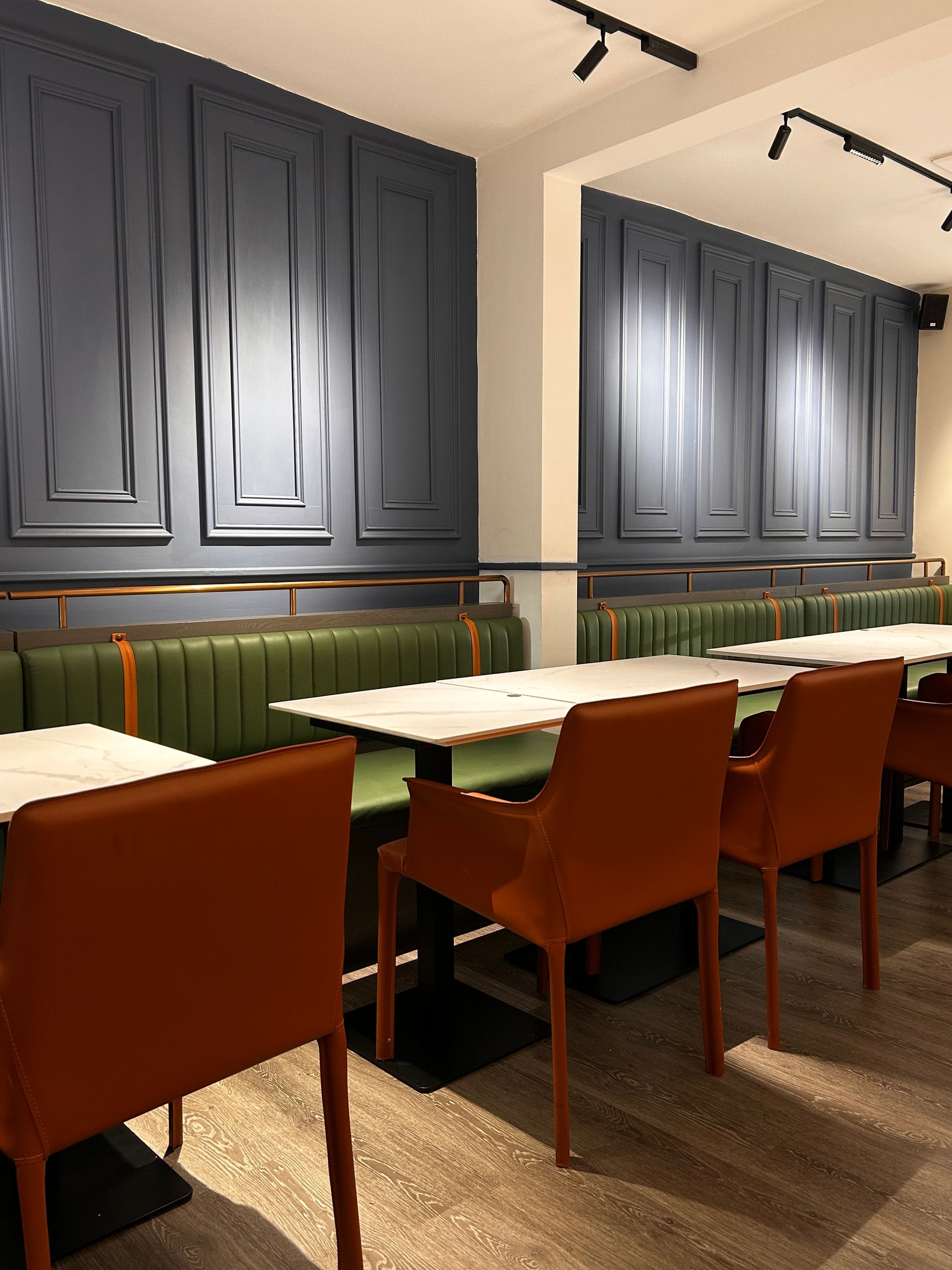 Restaurant interior with green booths, white tables, and orange chairs against blue paneling.