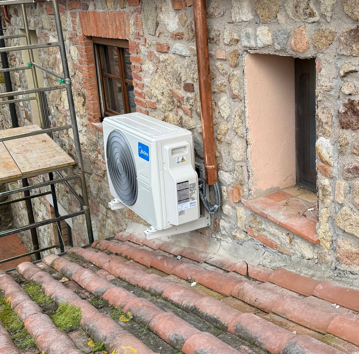 Un condizionatore d'aria è installato su un muro esterno in pietra, sopra un tetto di tegole in terracotta.