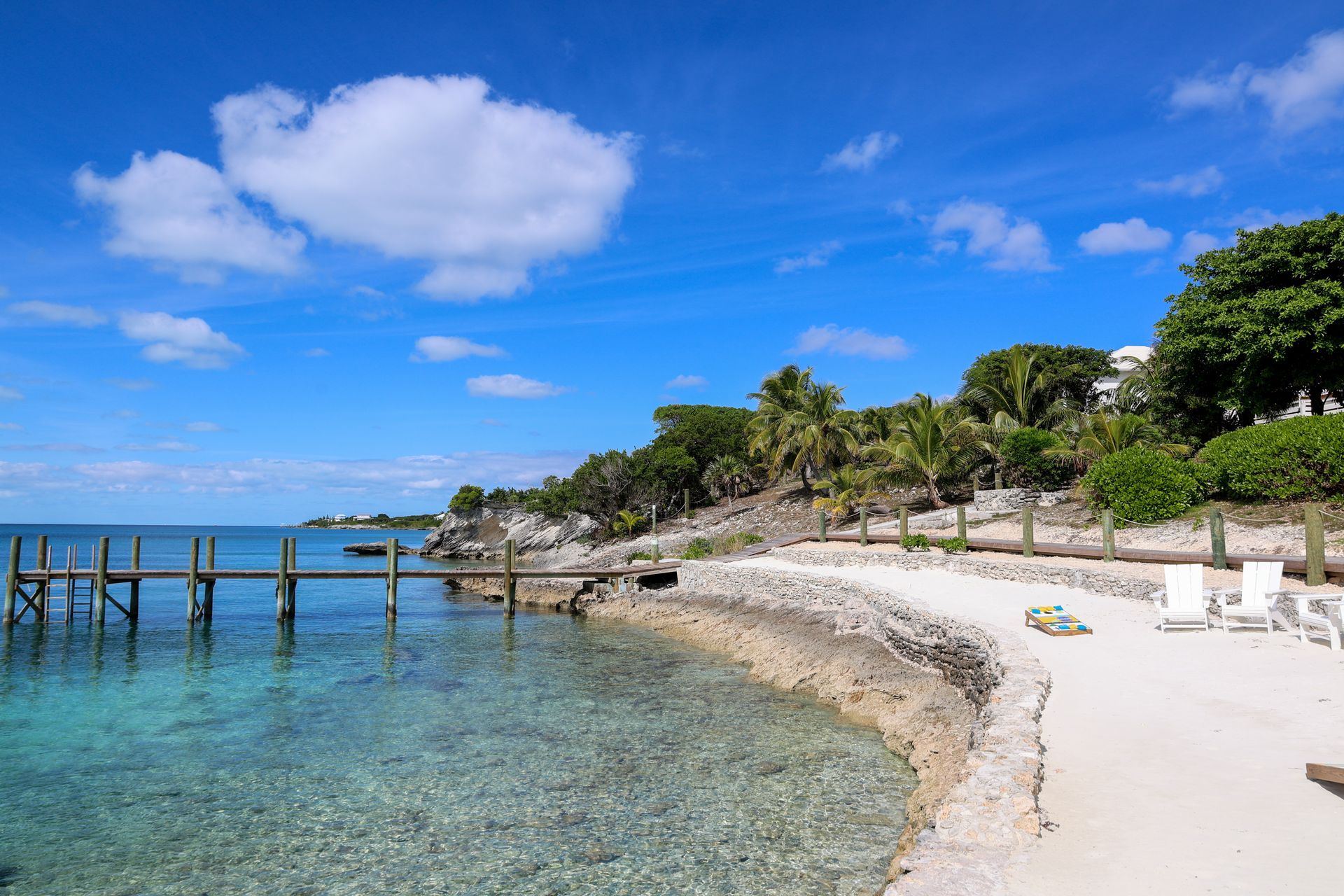 Sunny tropical beach with clear blue water, white sand, palm trees, and a wooden pier under a bright sky