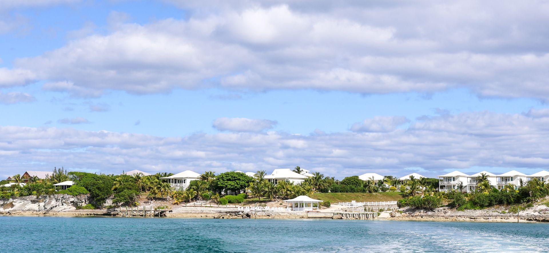Wide coastal view with blue water, rocky shore, palm trees, and white buildings under a partly cloudy sky