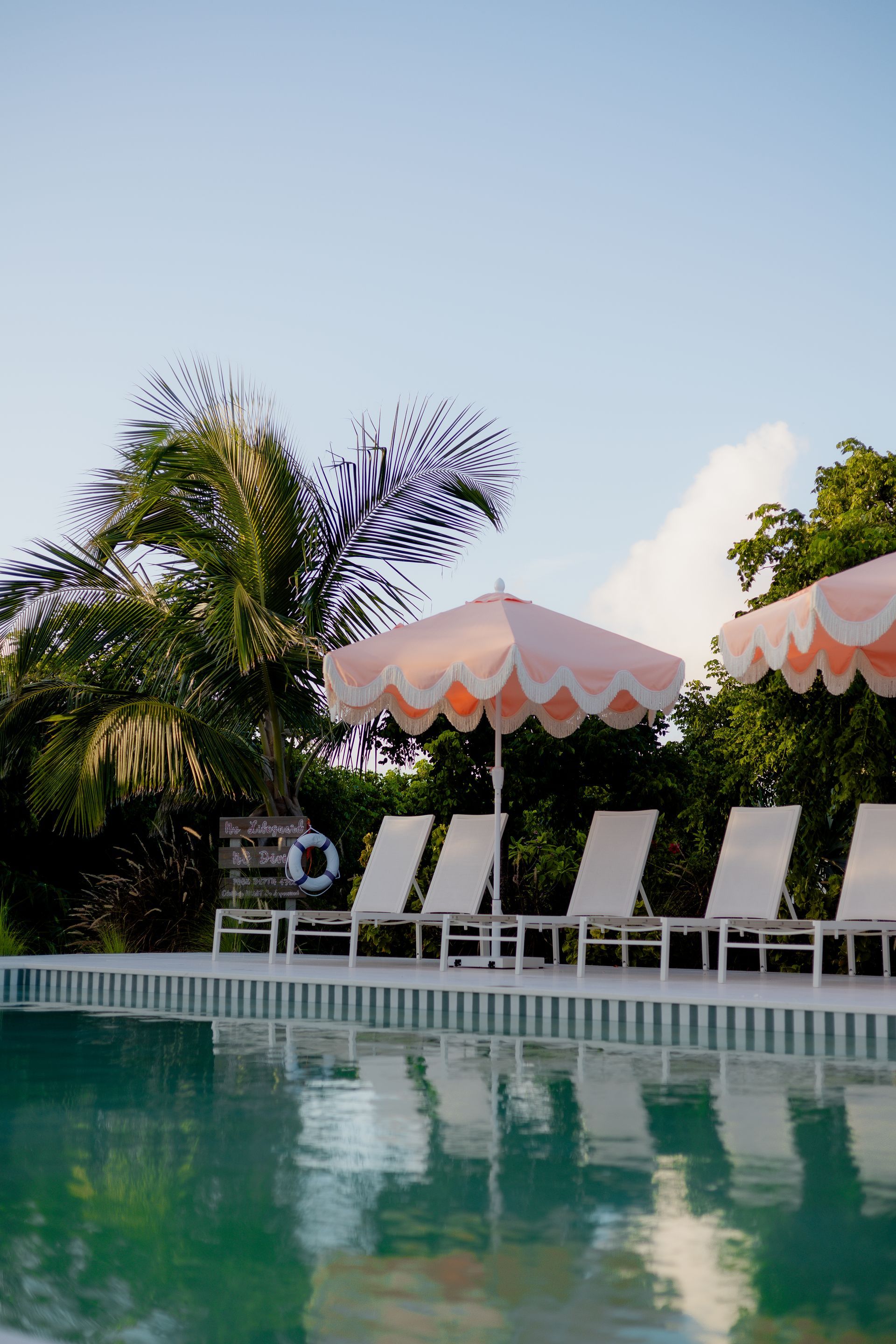 Resort pool with lounge chairs, striped umbrellas, palm trees, and calm water reflecting the scene